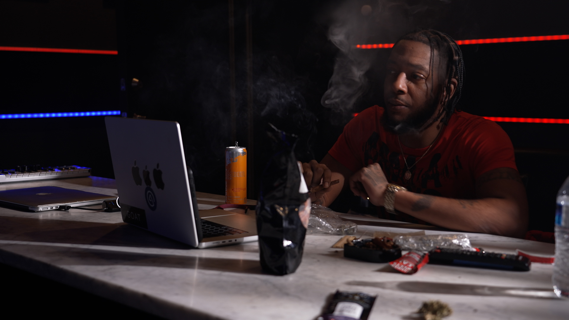 Person with braided hair sitting at a table with a laptop, smoking, with various items on the table.