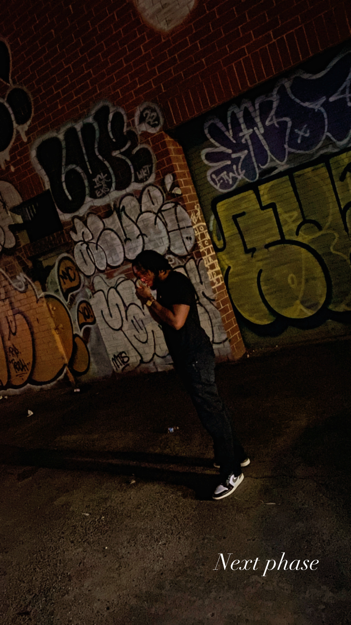 Person standing in front of graffiti-covered wall at night, holding a phone, with a shadow cast on the ground.