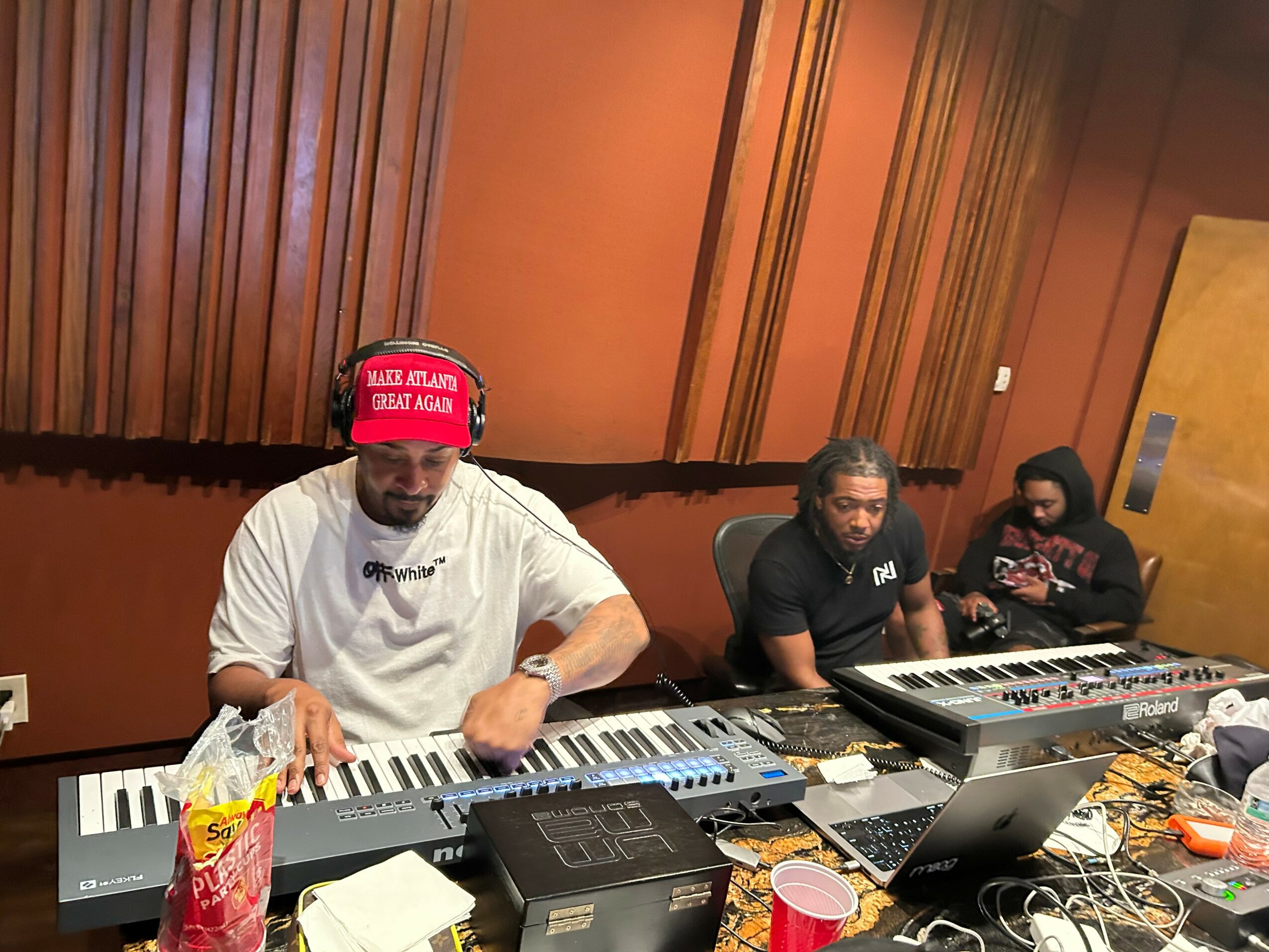 Three people sitting at a table with electronic music equipment, including keyboards and a laptop, in a room with wooden paneling.