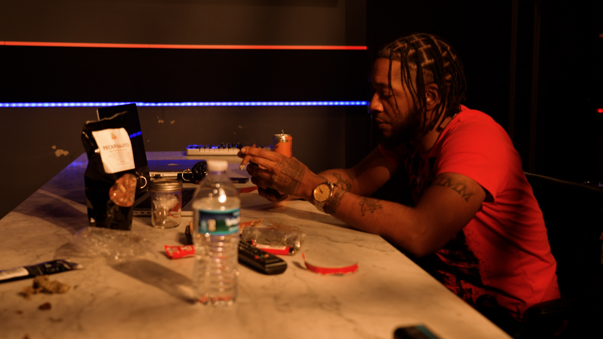 Person with braided hair and tattoos sitting at a table with drinks and electronic devices.