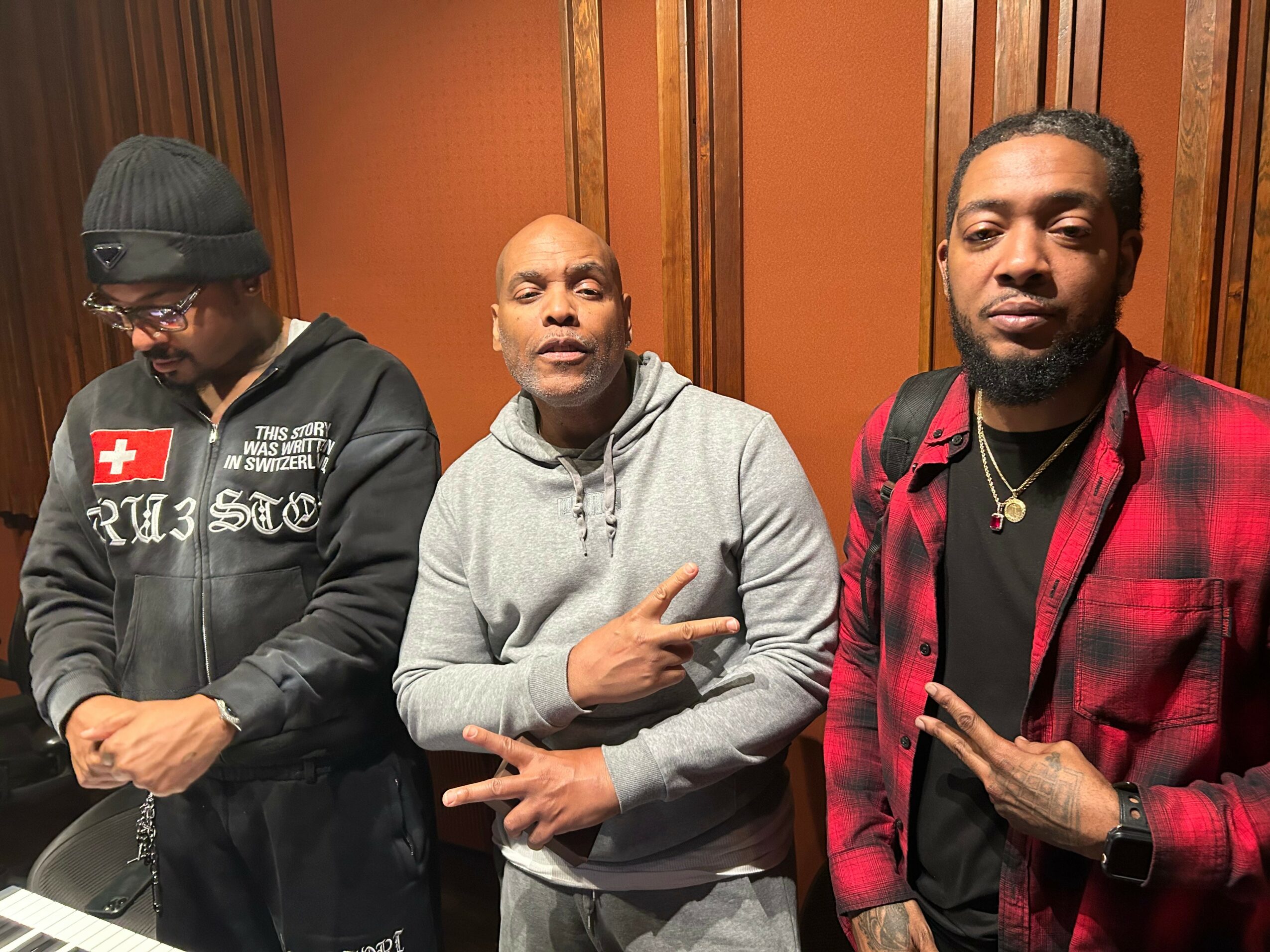 Three men standing indoors, one in a gray hoodie making a peace sign, another in a red plaid jacket, and the third in a black hoodie with a Swiss flag patch, all facing forward.