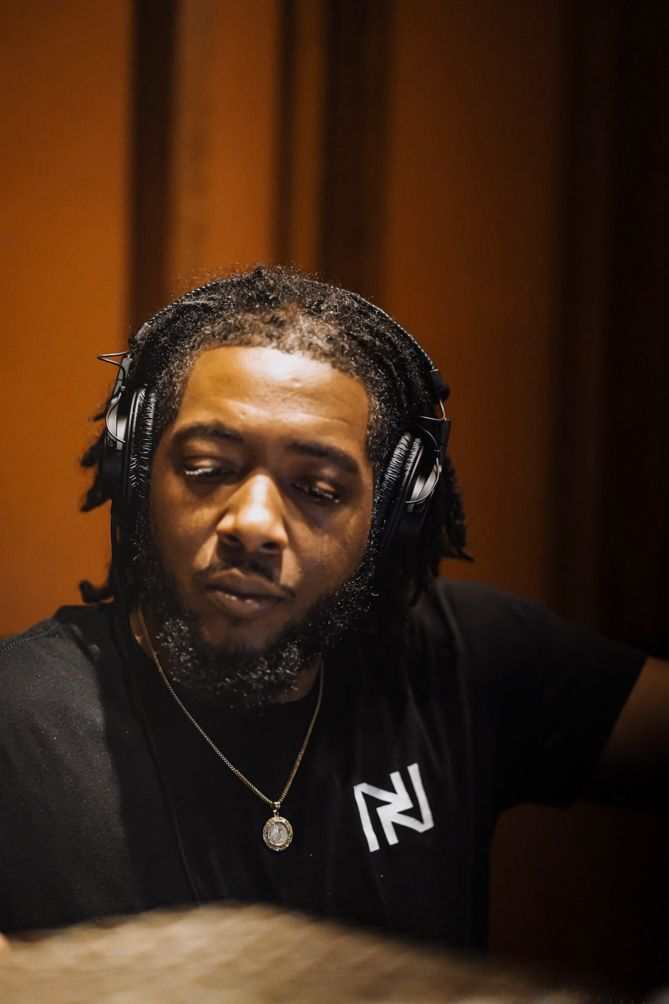 Man with dreadlocks wearing headphones and a black shirt with a logo, in a recording studio.