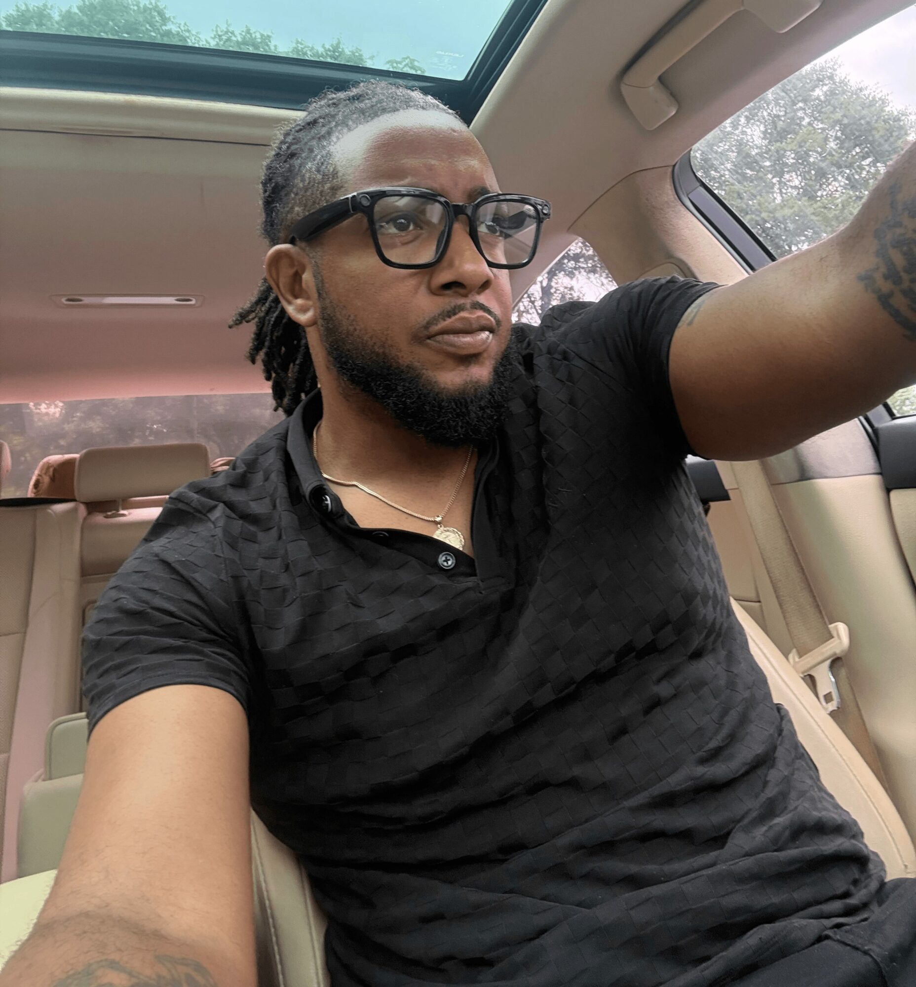 Man with glasses and dreadlocks taking a selfie inside a car, wearing a black shirt, with a sunroof and trees visible outside.
