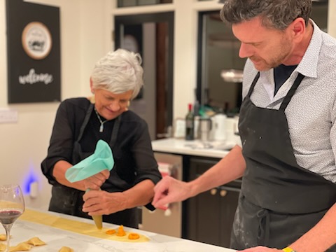 An elderly woman and a man are in a kitchen, preparing food together, smiling and holding hands over a yellow table.