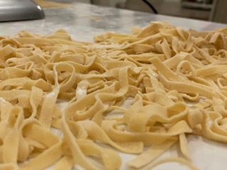 Uncooked pasta, likely fettuccine, spread out on a surface, with a kitchen background visible in the distance.