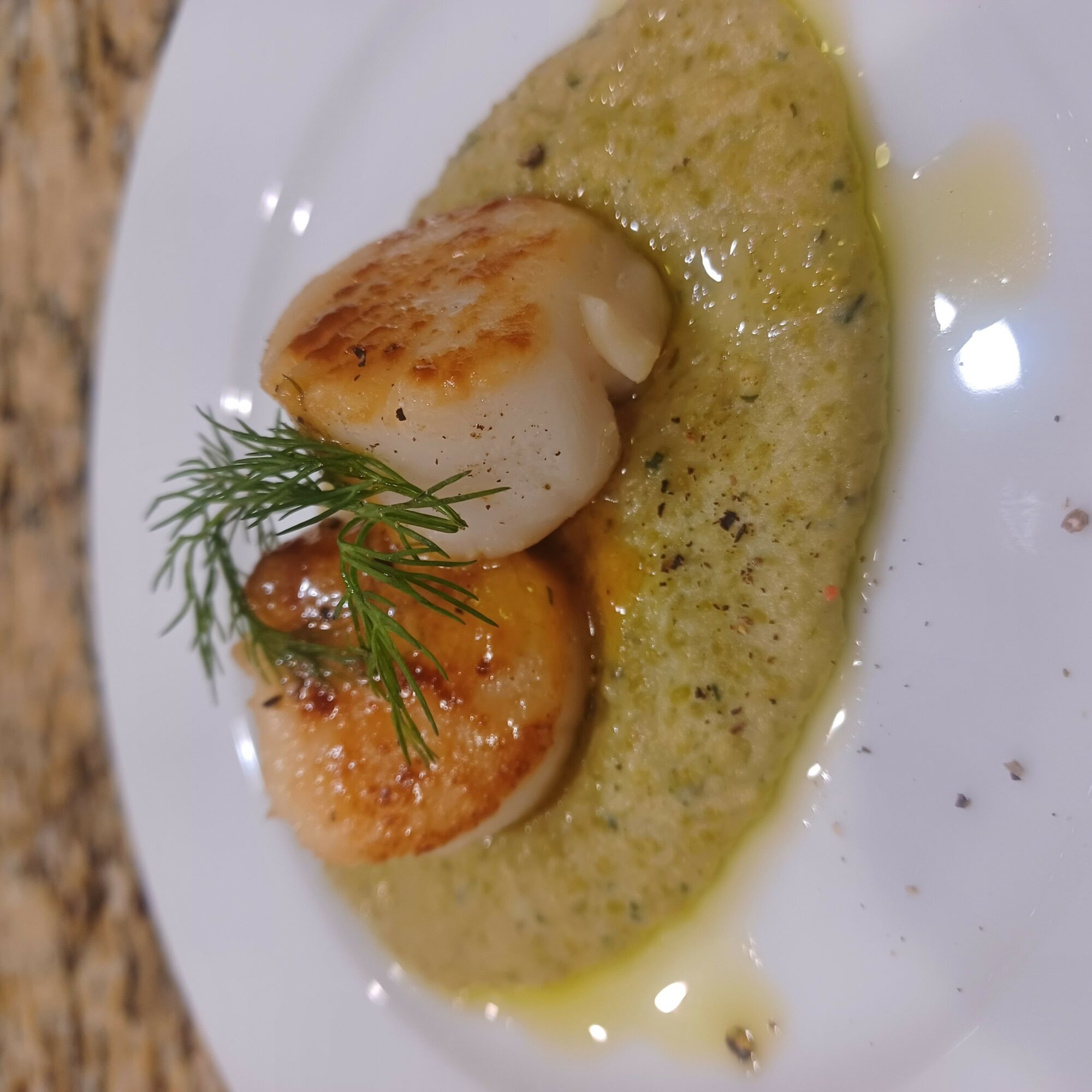 Two seared scallops garnished with dill, served on a green sauce on a white plate.