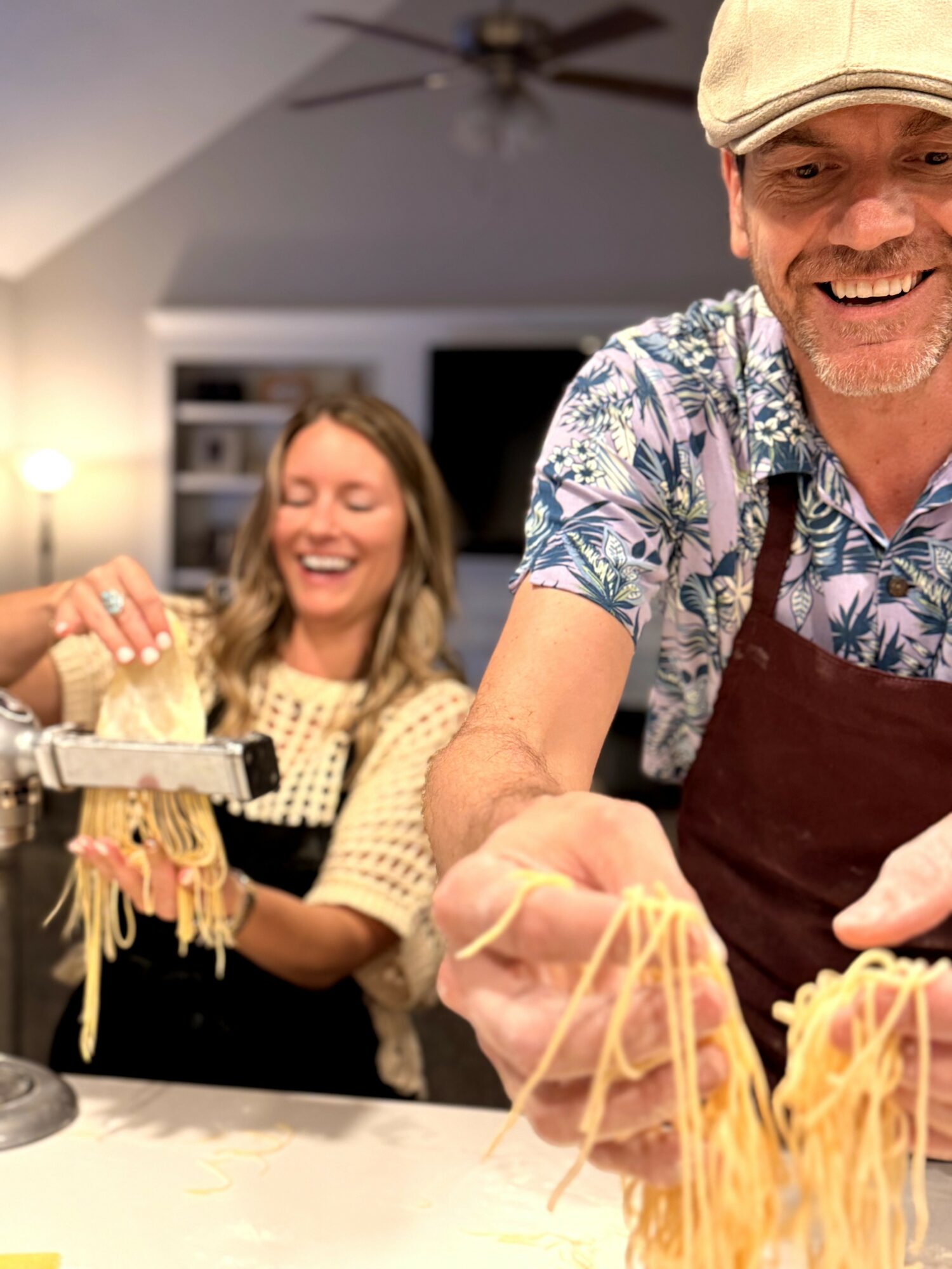 Two people preparing spaghetti in a kitchen, smiling and wearing casual clothes, with a ceiling fan overhead.
