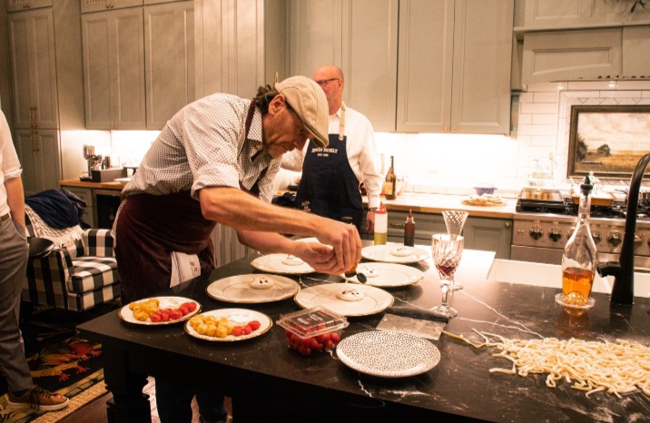 Person preparing food in a kitchen with plates and ingredients on a black countertop, others in the background, warm lighting.