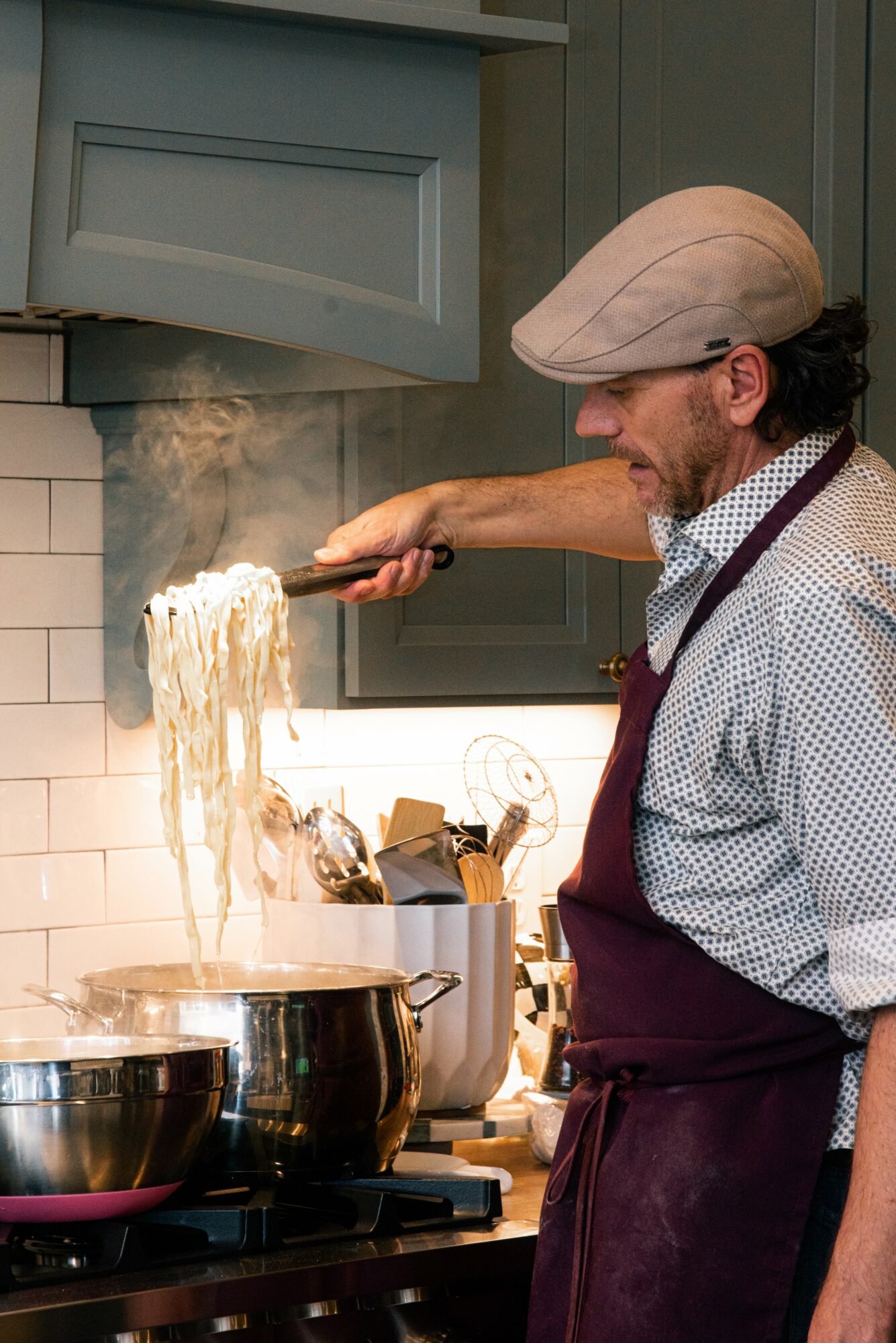 Man cooking pasta in a kitchen, wearing a flat cap and apron, stirring boiling water with steam rising.