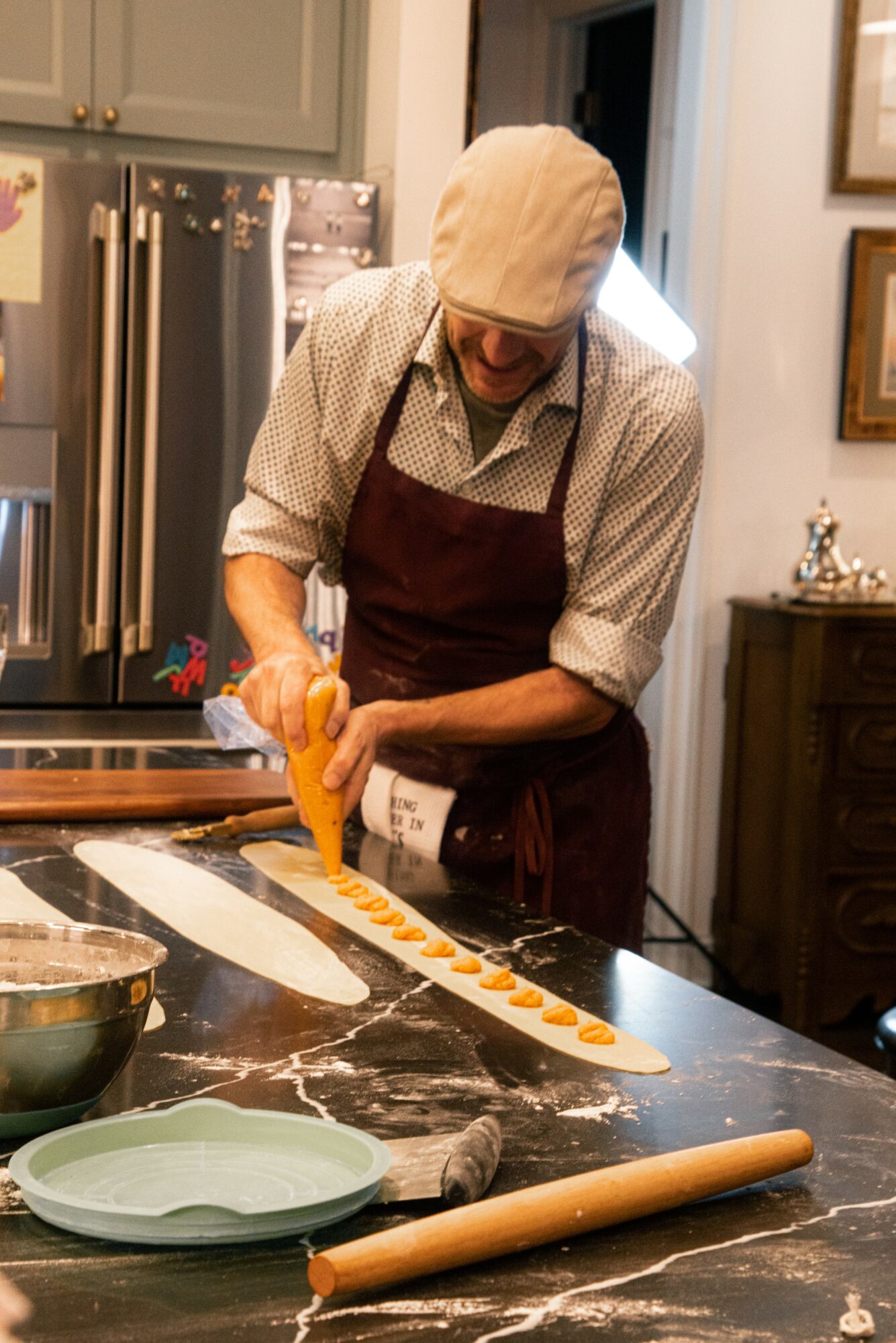 Person in apron and hat piping dough onto a marble countertop in a kitchen, with baking tools nearby.