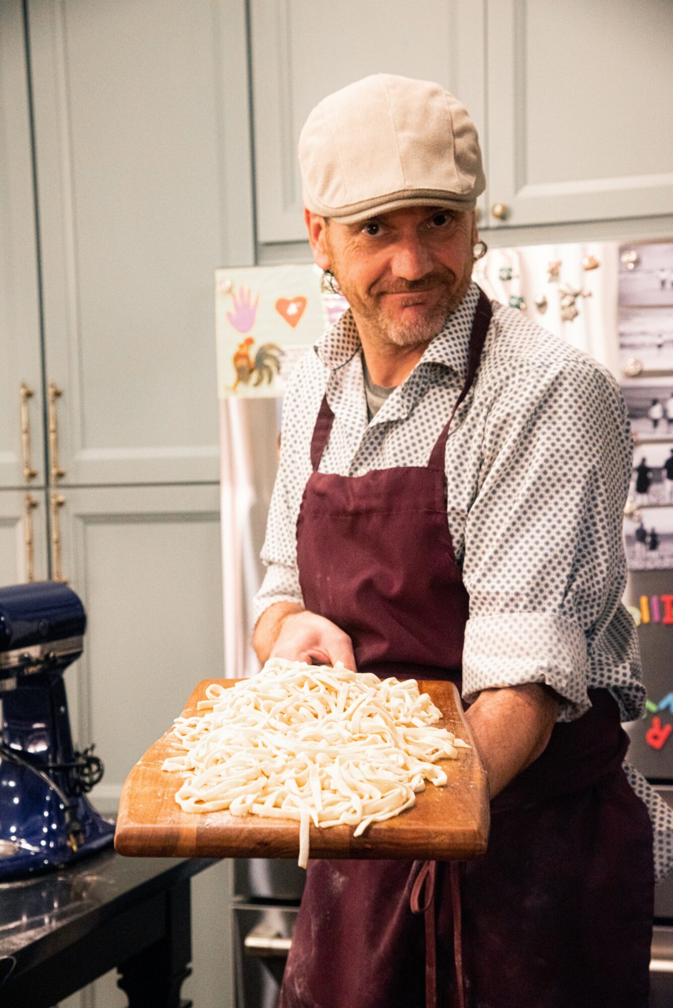 Person wearing a beige cap and apron holds a wooden board with spaghetti noodles in a kitchen.