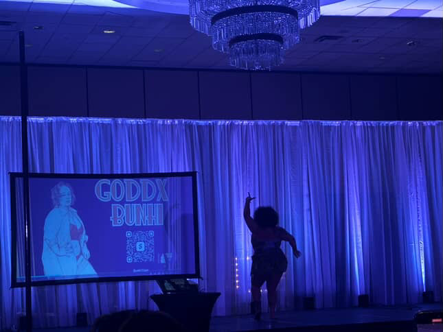 Child standing on stage with a raised arm, curtains and a screen in the background, blue lighting, chandelier overhead.