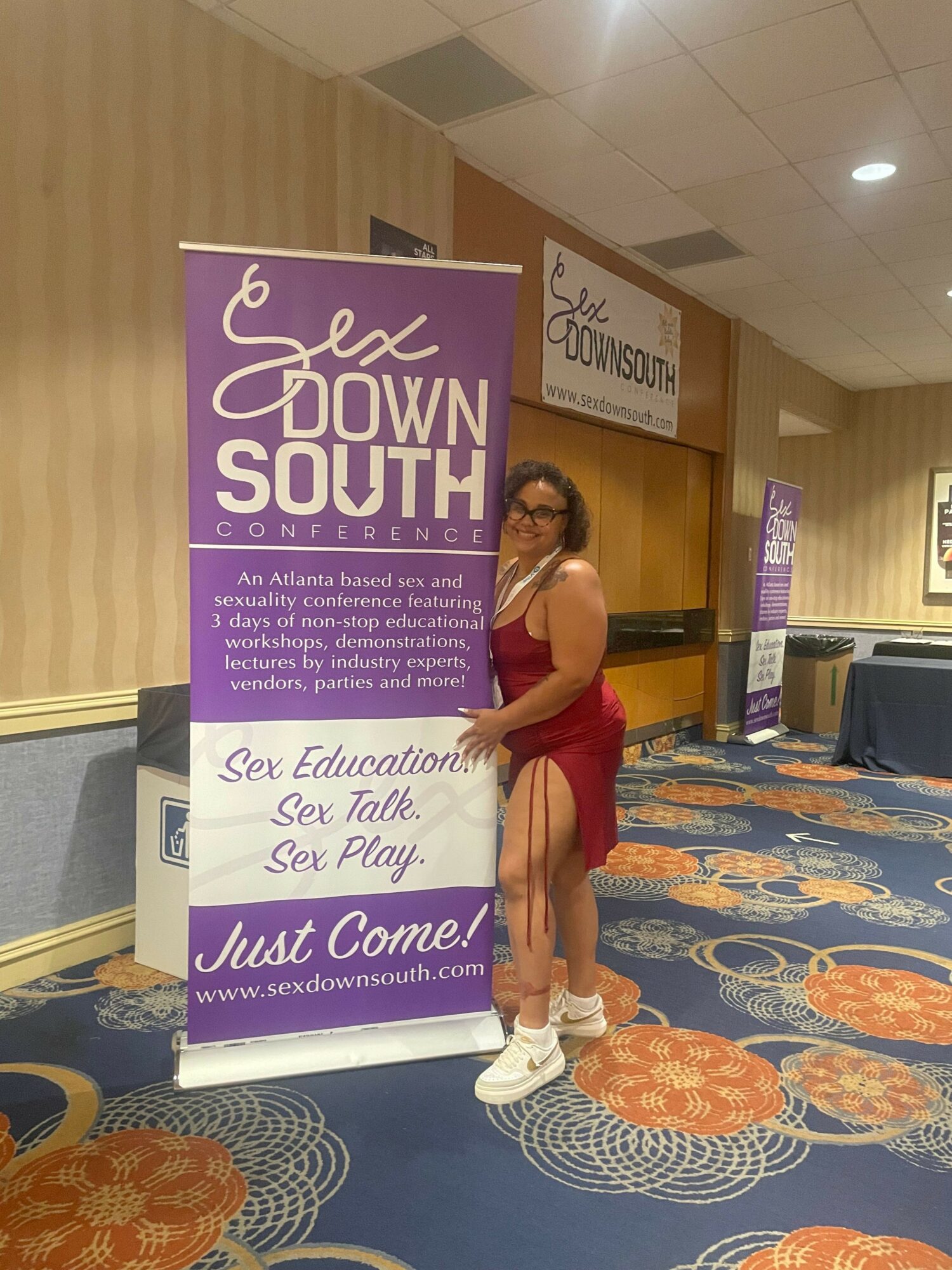 Child standing next to a purple and white informational sign at an indoor event, with a patterned carpet and a banner in the background.