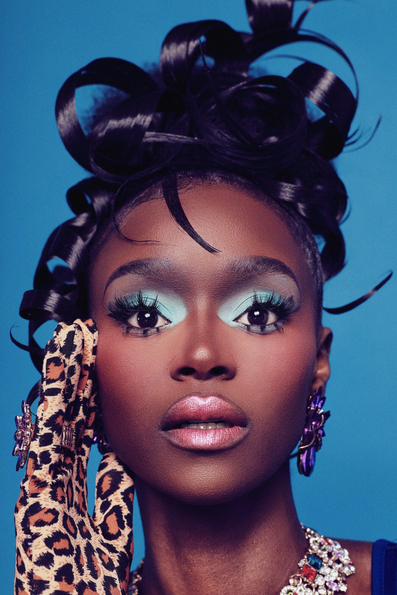 Portrait of a woman with elaborate hairstyle, bold makeup, and leopard print accessory, against a blue background.