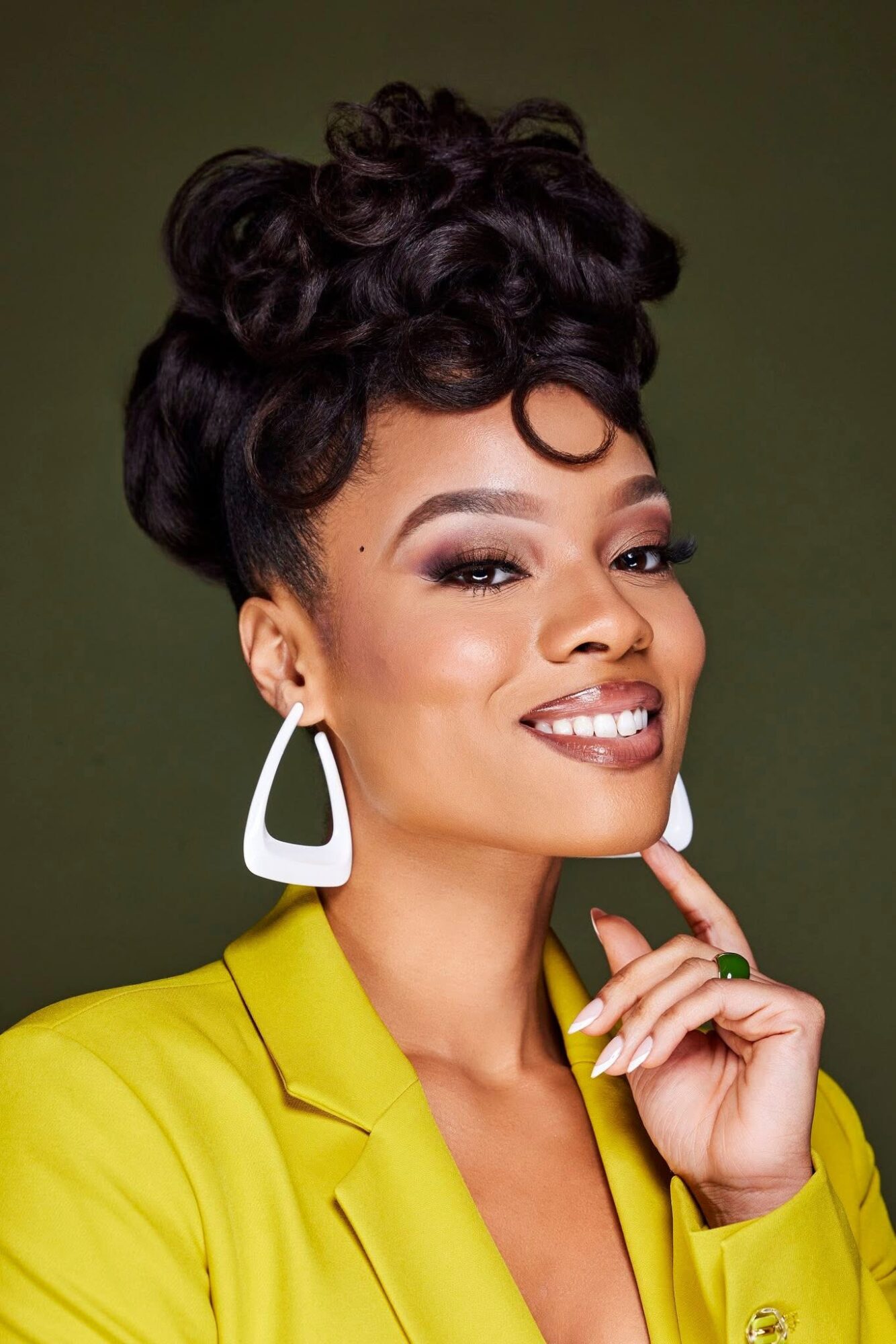 Woman with styled hair, large earrings, yellow blazer, smiling, touching her chin, against a dark background.