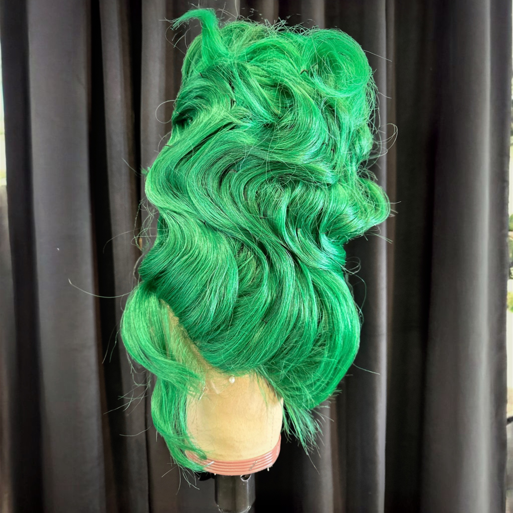 Green wig with large curls on a mannequin head against black curtains.