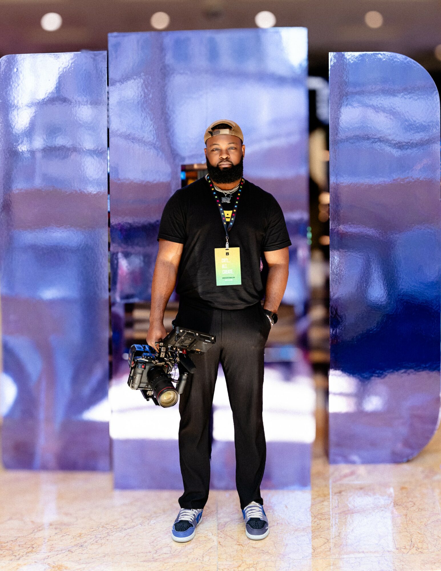 Man with a beard and glasses stands indoors holding a camera, wearing a black shirt, pants, and sneakers, with a lanyard.