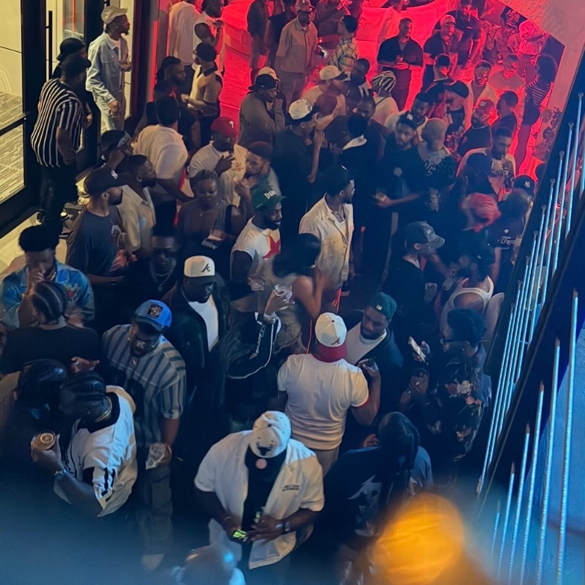 Crowd of people gathered indoors with red and blue lighting, some wearing hats and casual clothing.