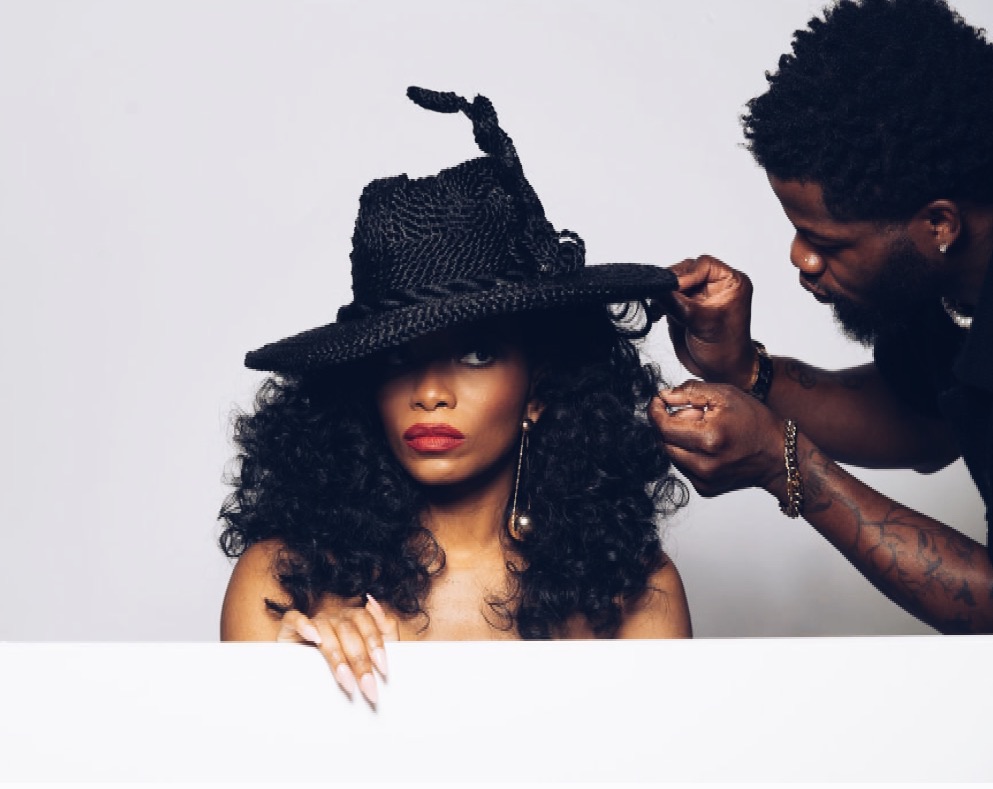 A woman with curly hair wears a large black hat, while a man adjusts it. She looks at the camera.