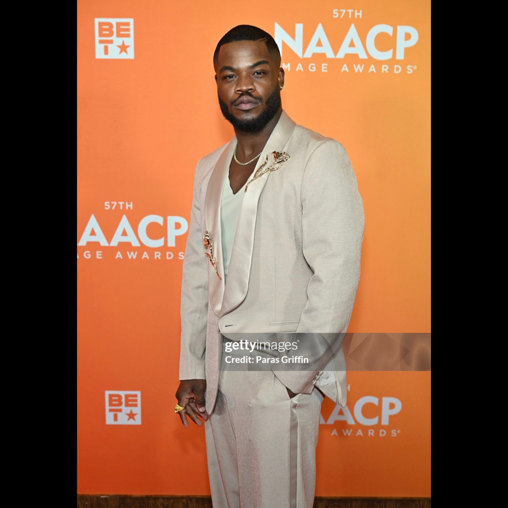 Man in light-colored suit standing in front of orange backdrop with event logos, looking at camera.