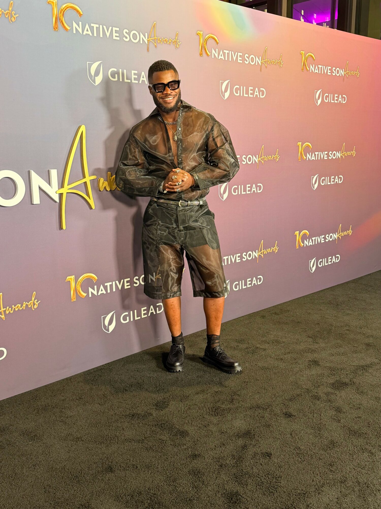 Person standing on stage with a purple backdrop, holding a microphone, wearing a leather jacket and shorts.