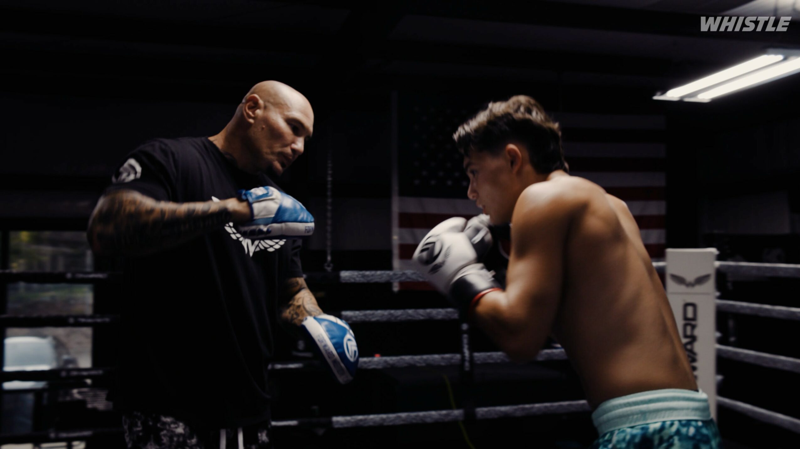 Boxer and trainer in a boxing ring, trainer holding gloves, boxer in stance, dimly lit gym environment.