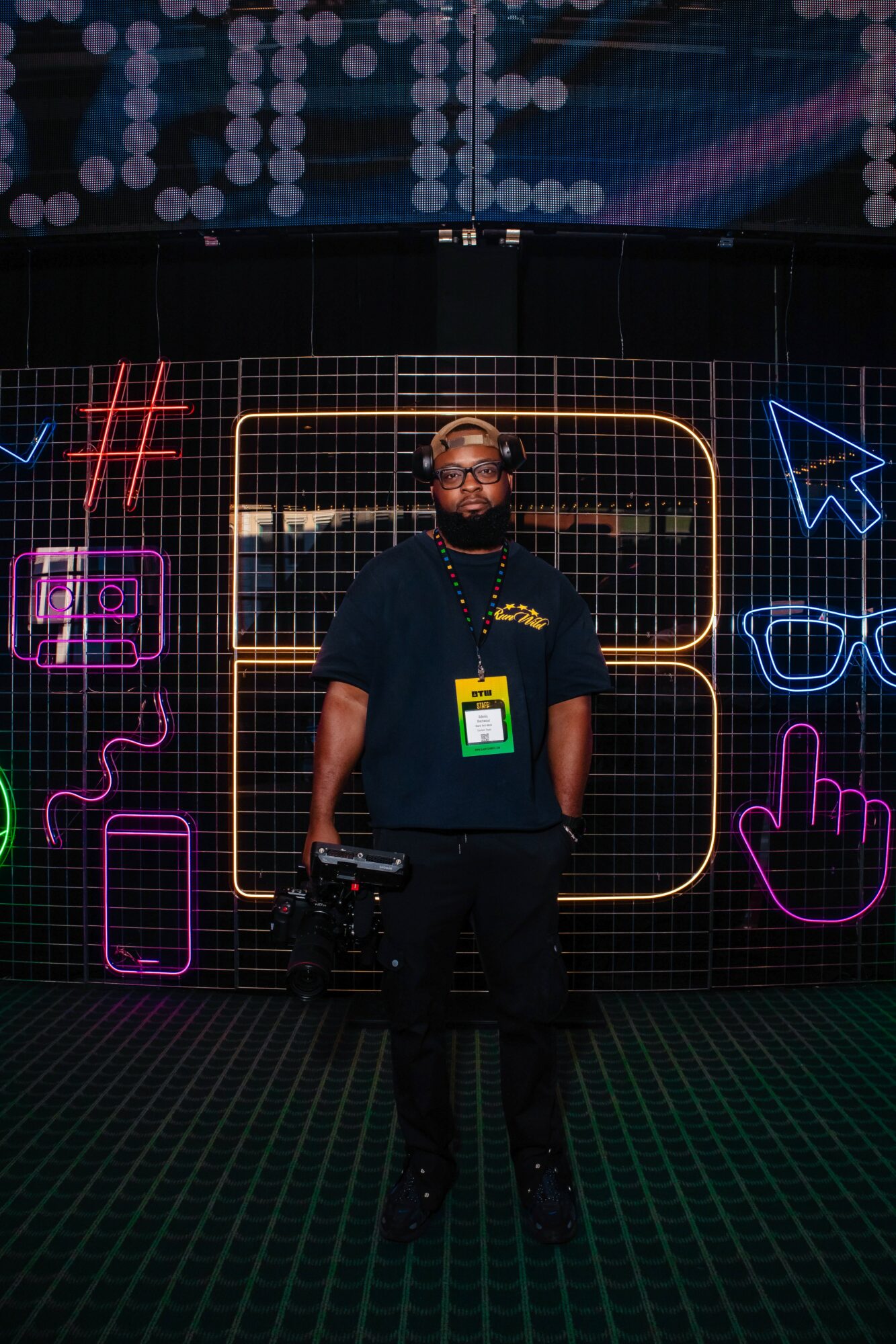 Person standing in front of colorful neon signs, wearing a black shirt, glasses, and a lanyard, with a camera hanging from their shoulder.