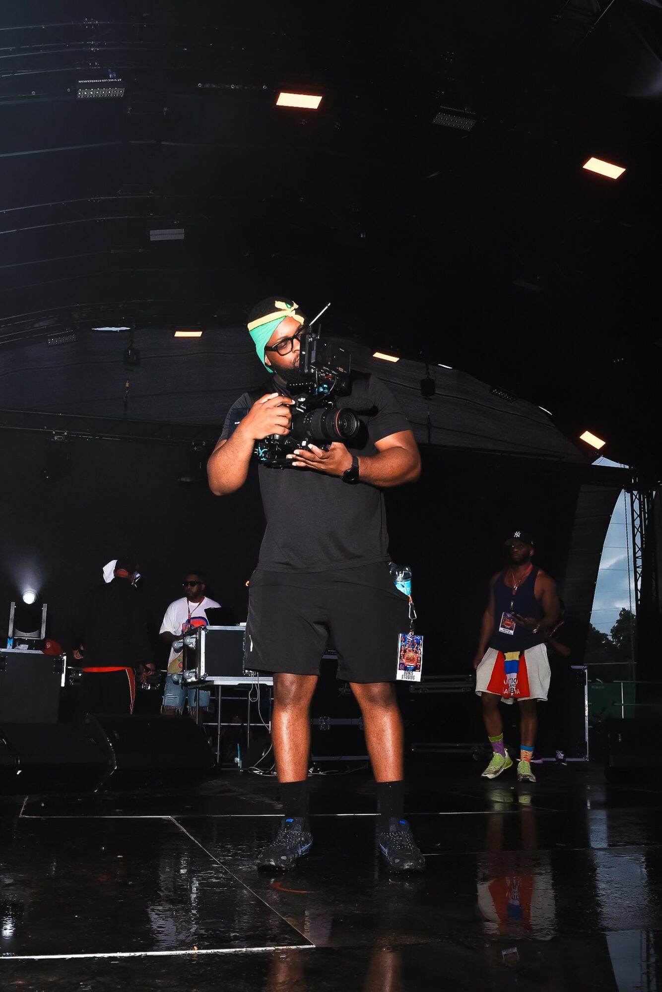 Person holding camera on stage with performers in background, dark setting with stage lights, reflective floor, and equipment.
