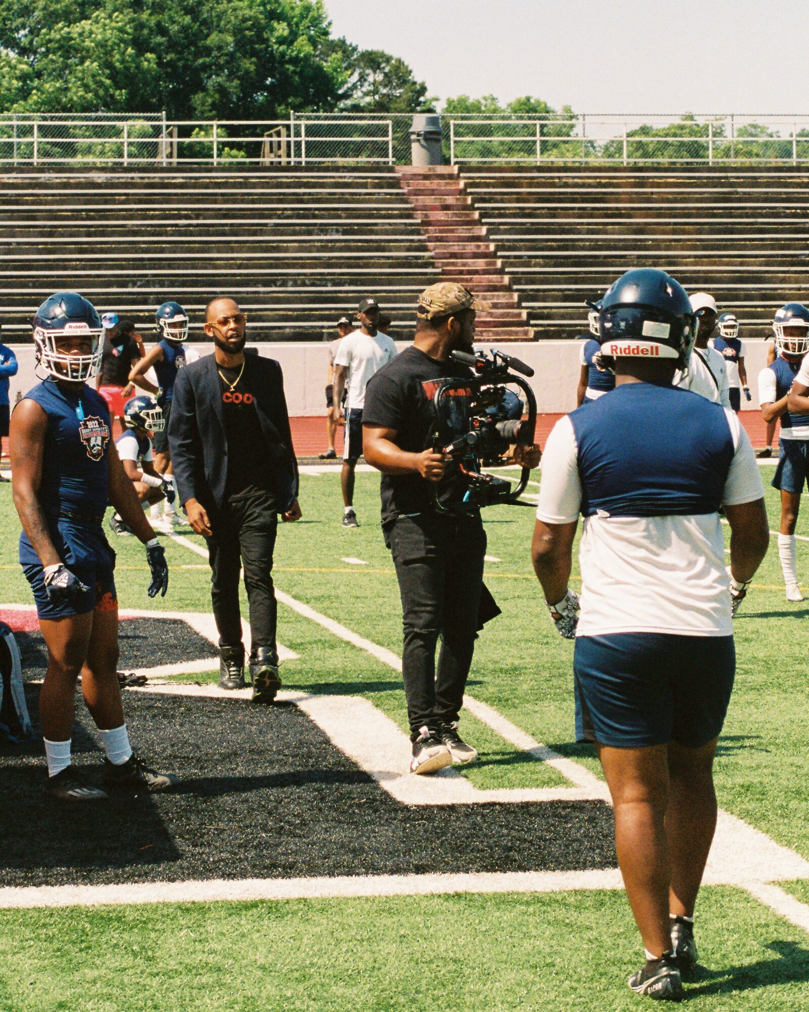 People on a football field, some wearing helmets, with a cameraman filming, and a man in a black jacket walking nearby.