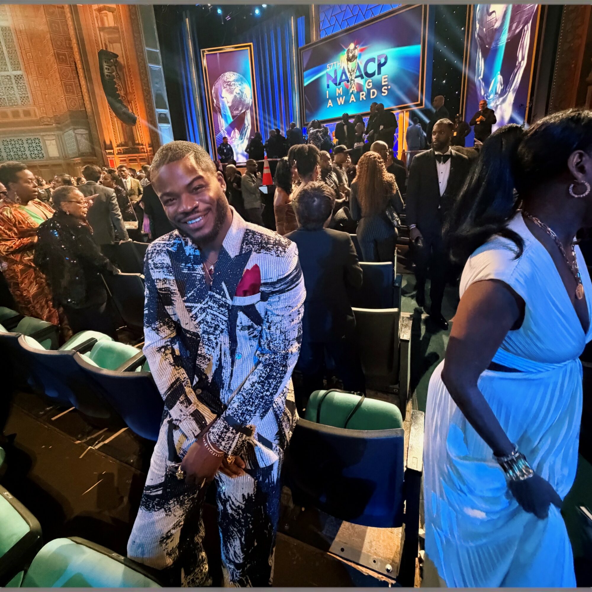Man in patterned suit smiling at an awards event, surrounded by seated and standing people, stage in background.