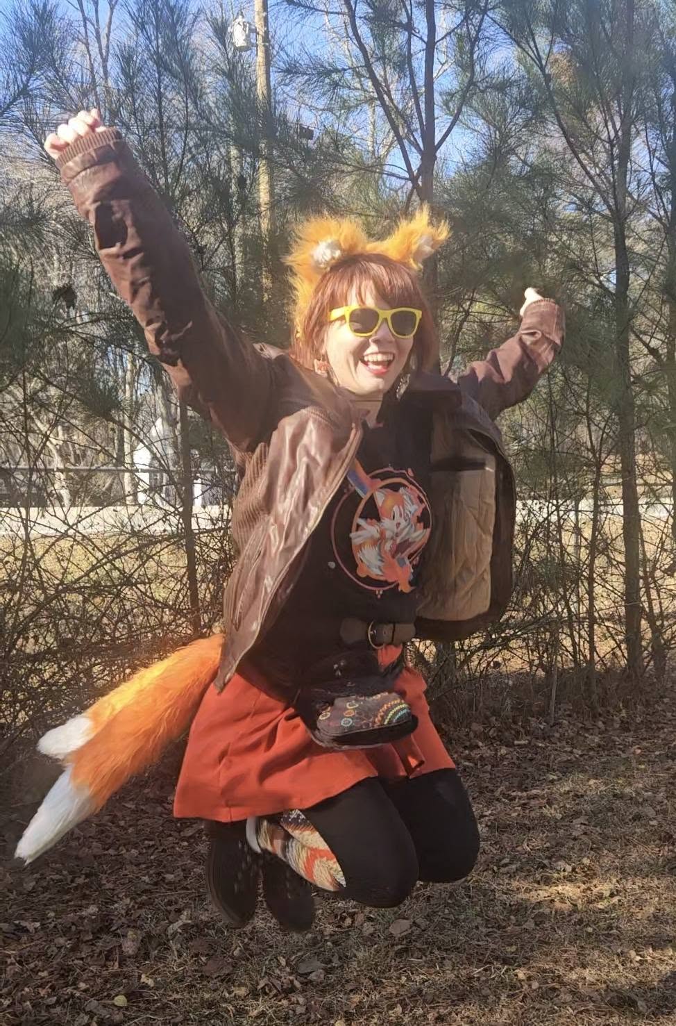 Person outdoors in a fox costume with orange tail, yellow sunglasses, jumping with arms raised, trees in background.
