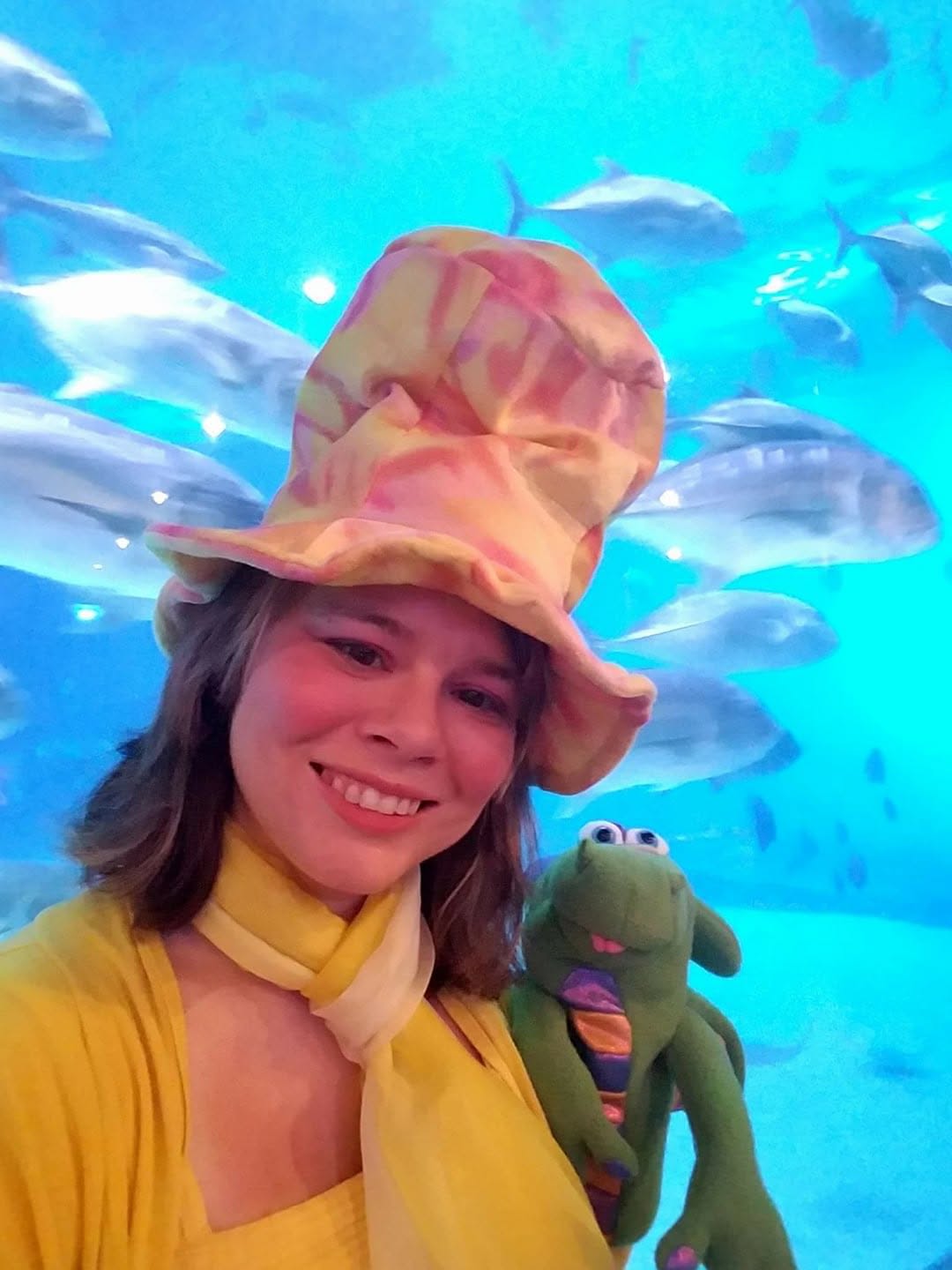 Girl wearing a colorful hat and yellow outfit, holding a plush toy, in front of an aquarium with fish.