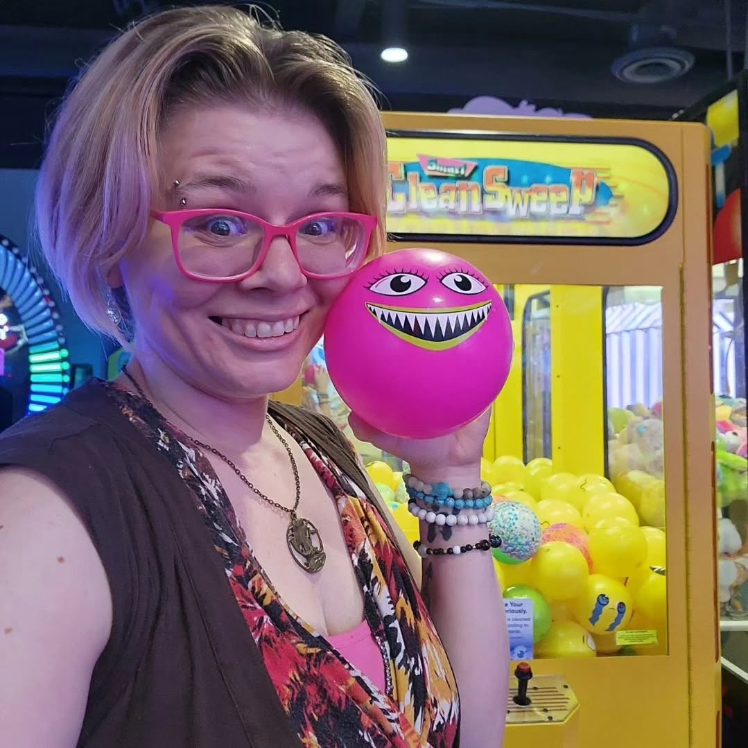 Woman with pink glasses smiling, holding a pink ball with a monster face, in arcade with claw machine in background.