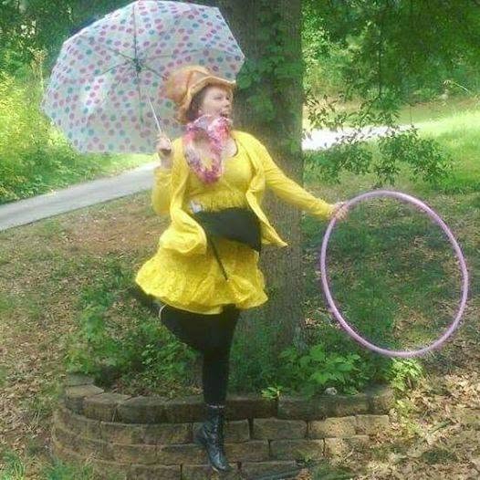 Person in yellow coat holding an umbrella and a hula hoop outdoors, standing on a brick border, surrounded by greenery.