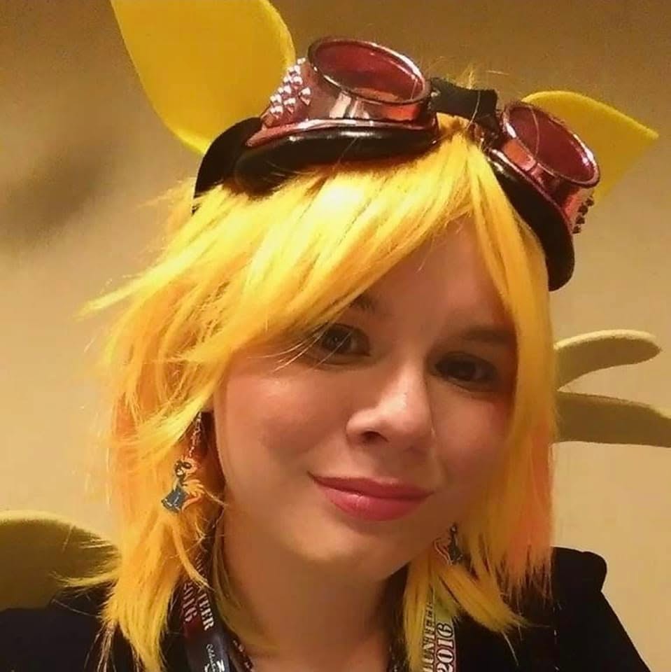 Person with yellow hair wearing goggles and yellow accessories, smiling, indoors.