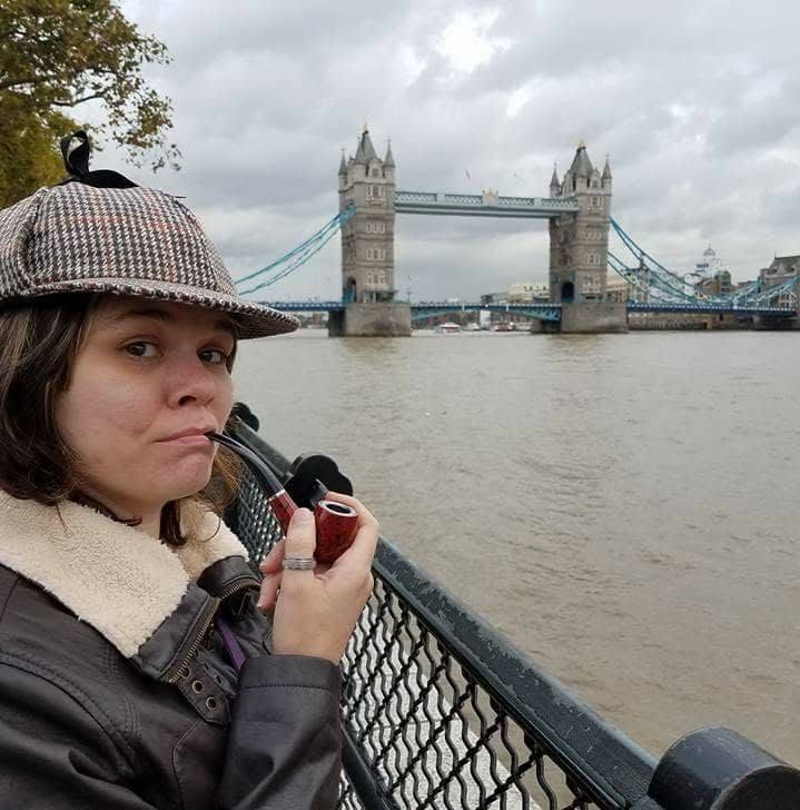Woman with a pipe and a checkered hat stands by the river with Tower Bridge in the background under cloudy sky.