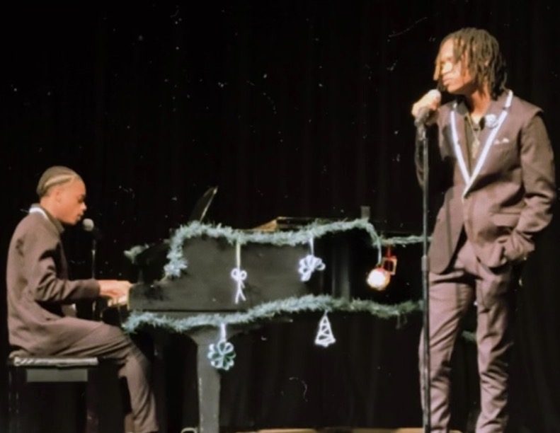 Two people on stage, one playing piano and the other singing into a microphone, with Christmas decorations on the piano.
