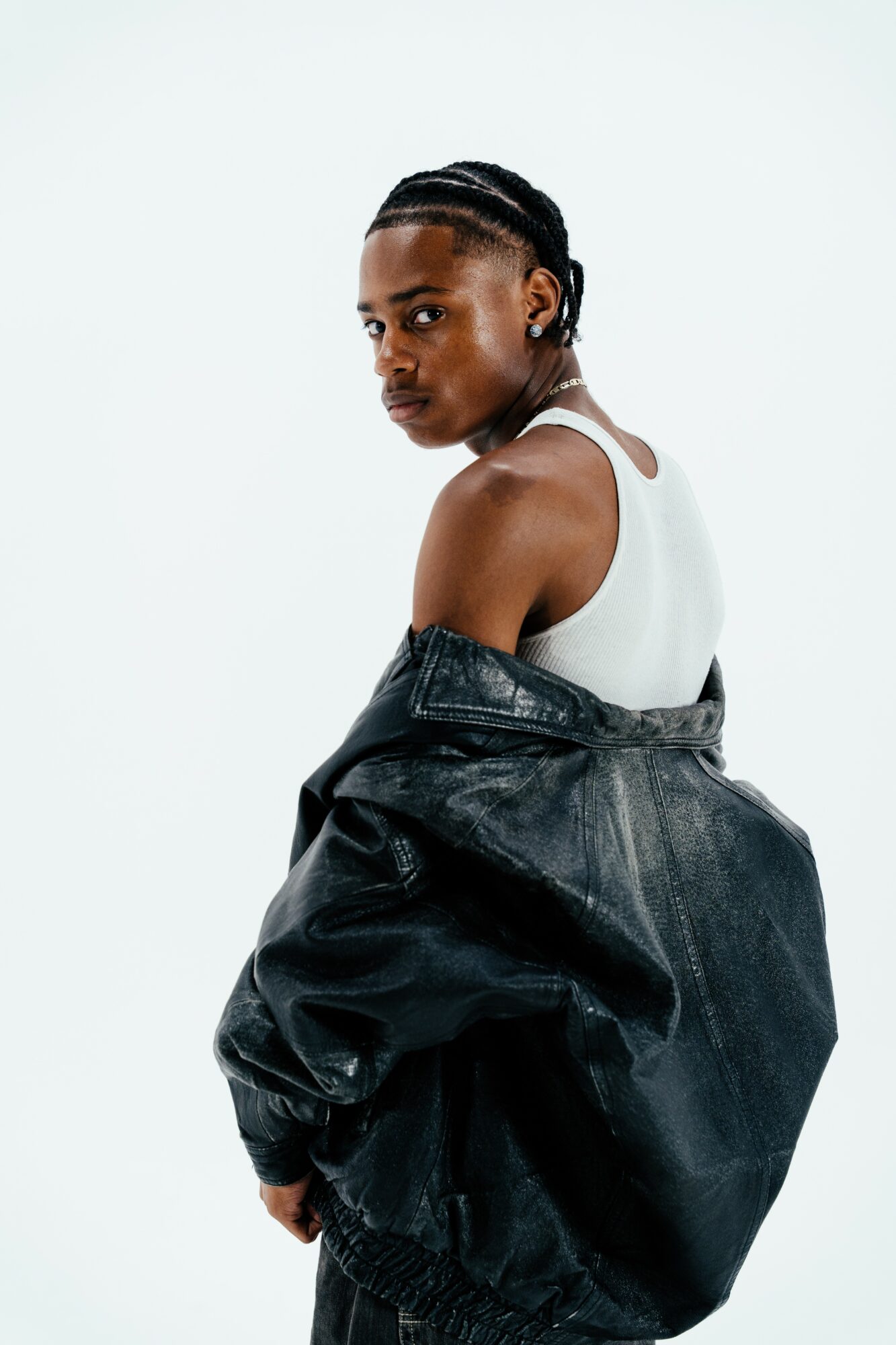 Young woman with braided hair wearing a white tank top and black jacket, looking over her shoulder against a plain background.