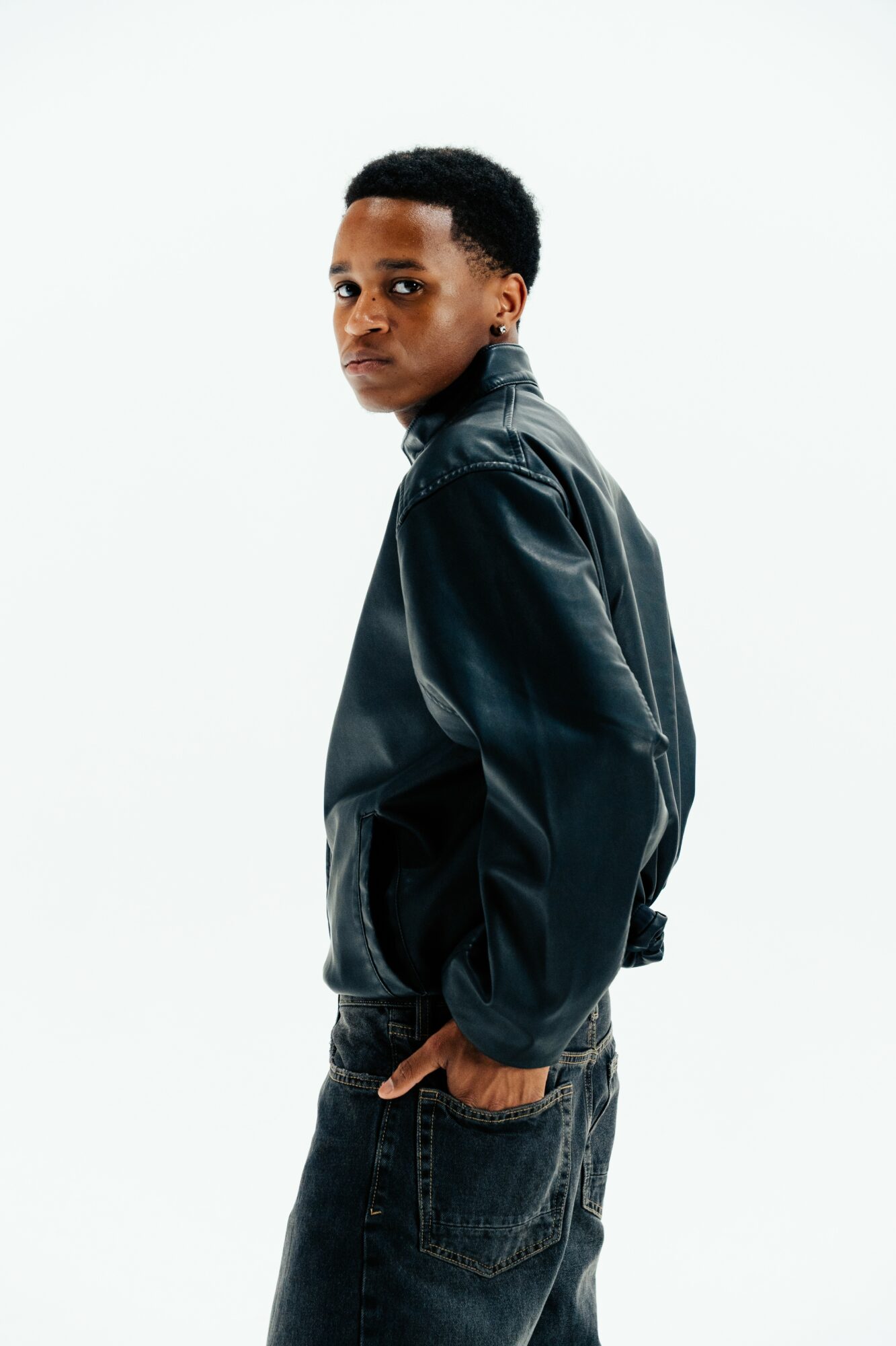 Young person with short hair wearing a black leather jacket and jeans, looking over shoulder against white background.