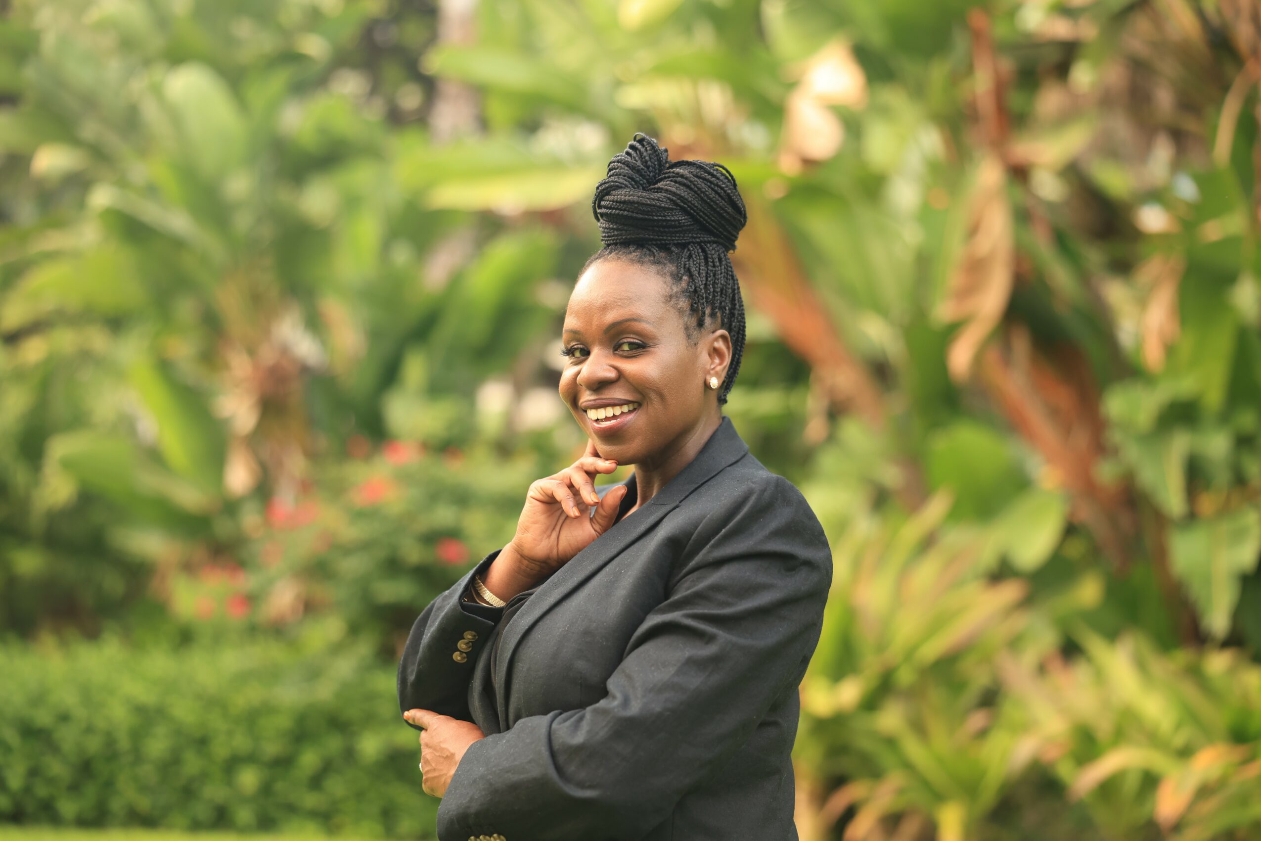 Smiling woman with braided hair in a bun, wearing a dark blazer, standing outdoors with green foliage background.