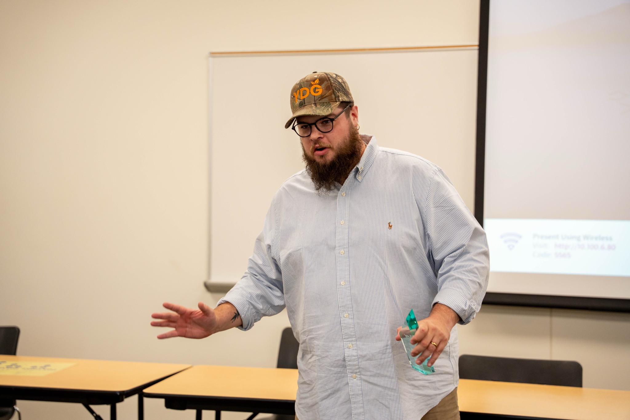 Man with glasses, beard, wearing a cap and light-colored shirt, standing and gesturing with his hands in a room with a presentation screen behind him.
