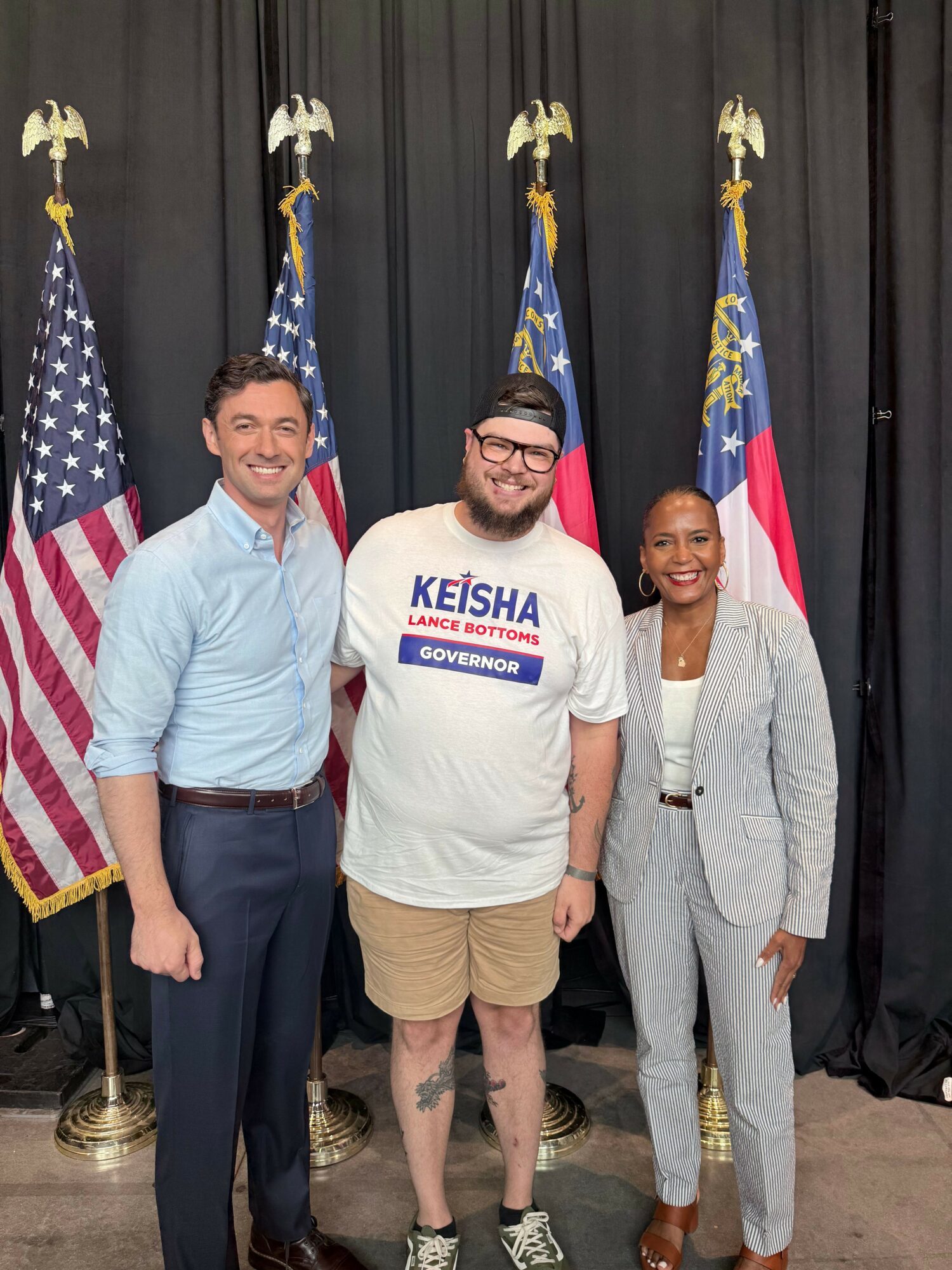 Three people standing in front of a black curtain with American flags, smiling, one wearing a KEISHA shirt, two in casual clothing.