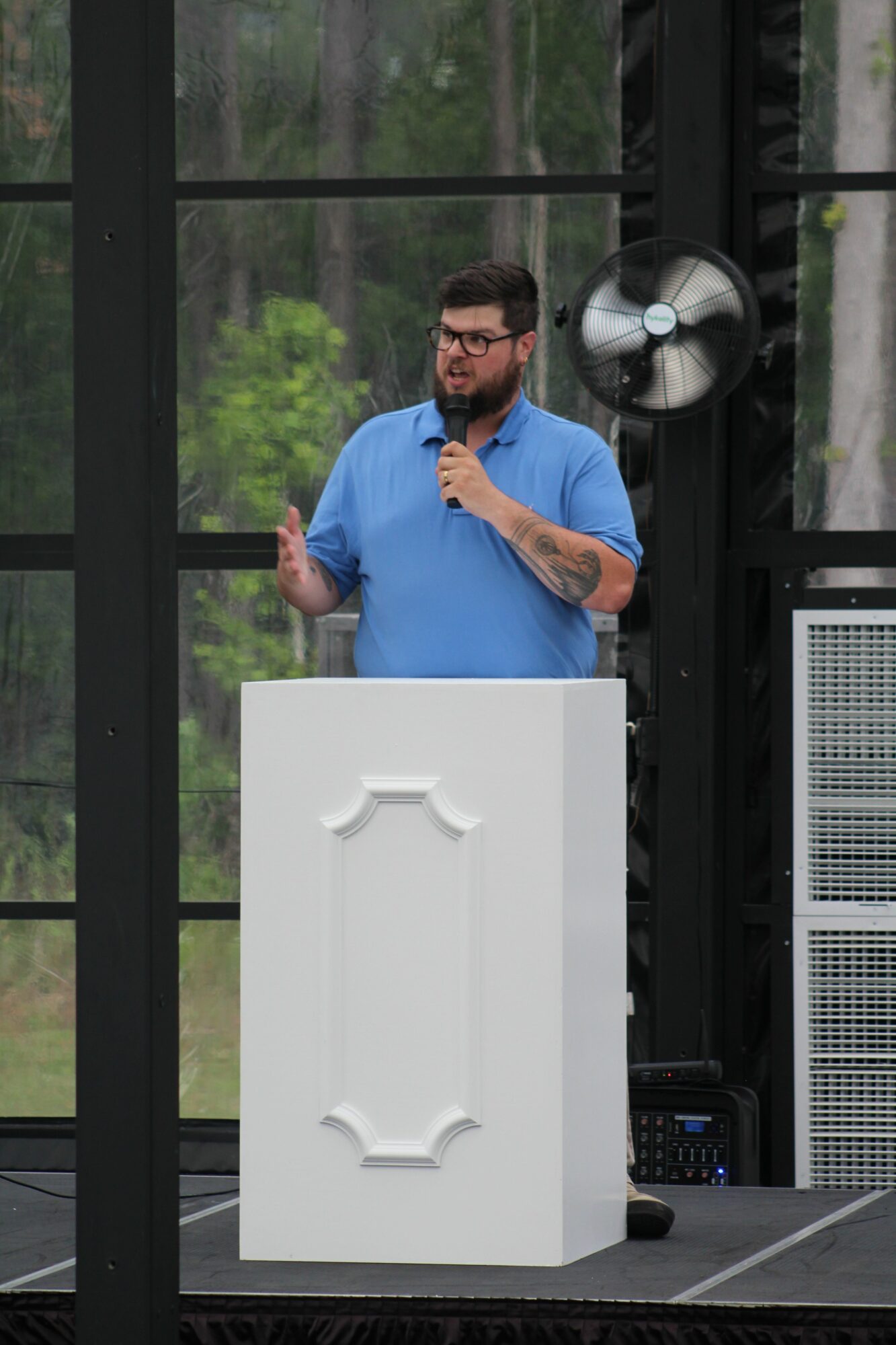 Man with glasses and beard in blue shirt standing behind a white podium, with a large fan and window in background.