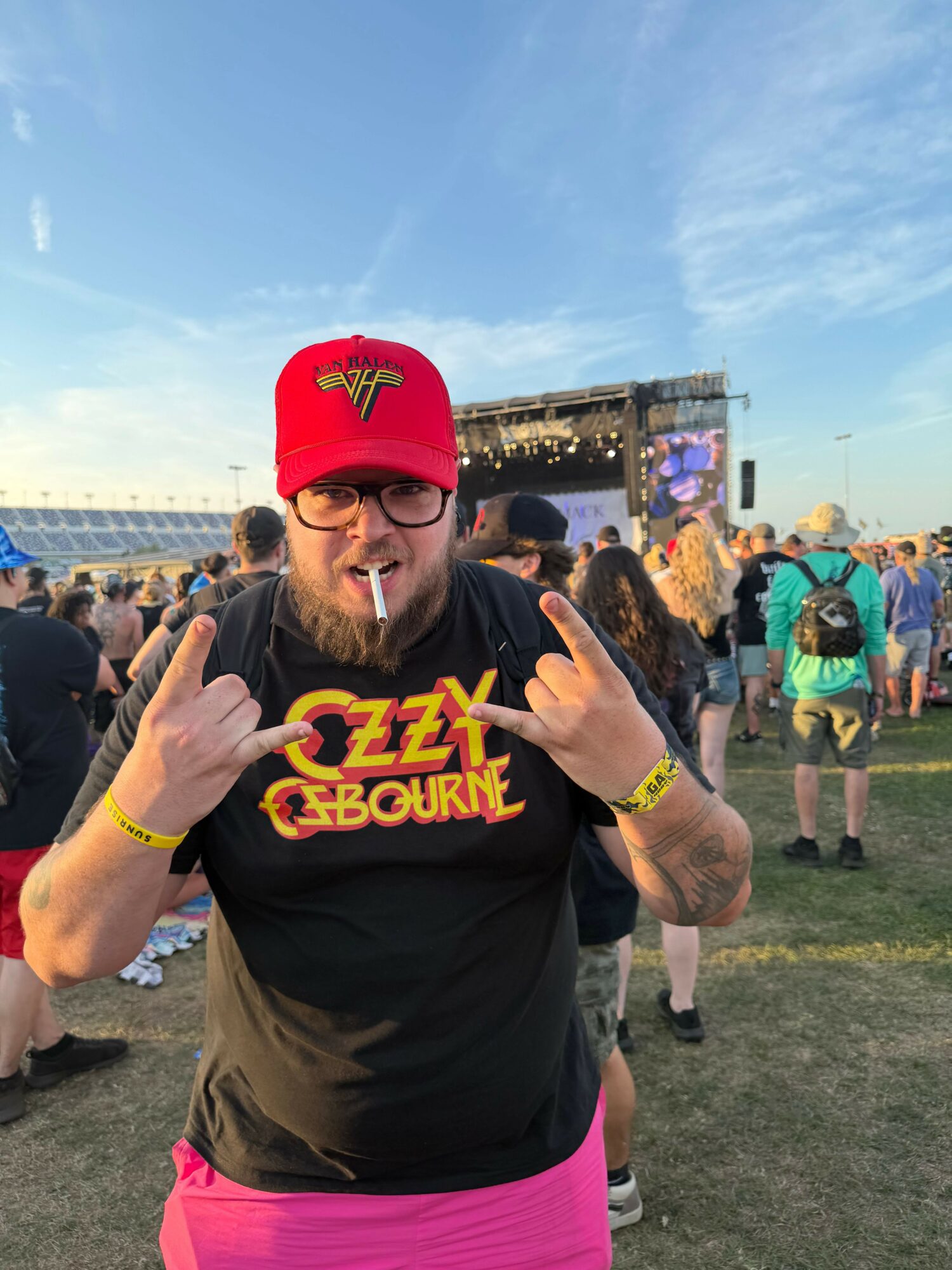 Person with a red cap, sunglasses, and a black T-shirt giving two thumbs up at an outdoor event.