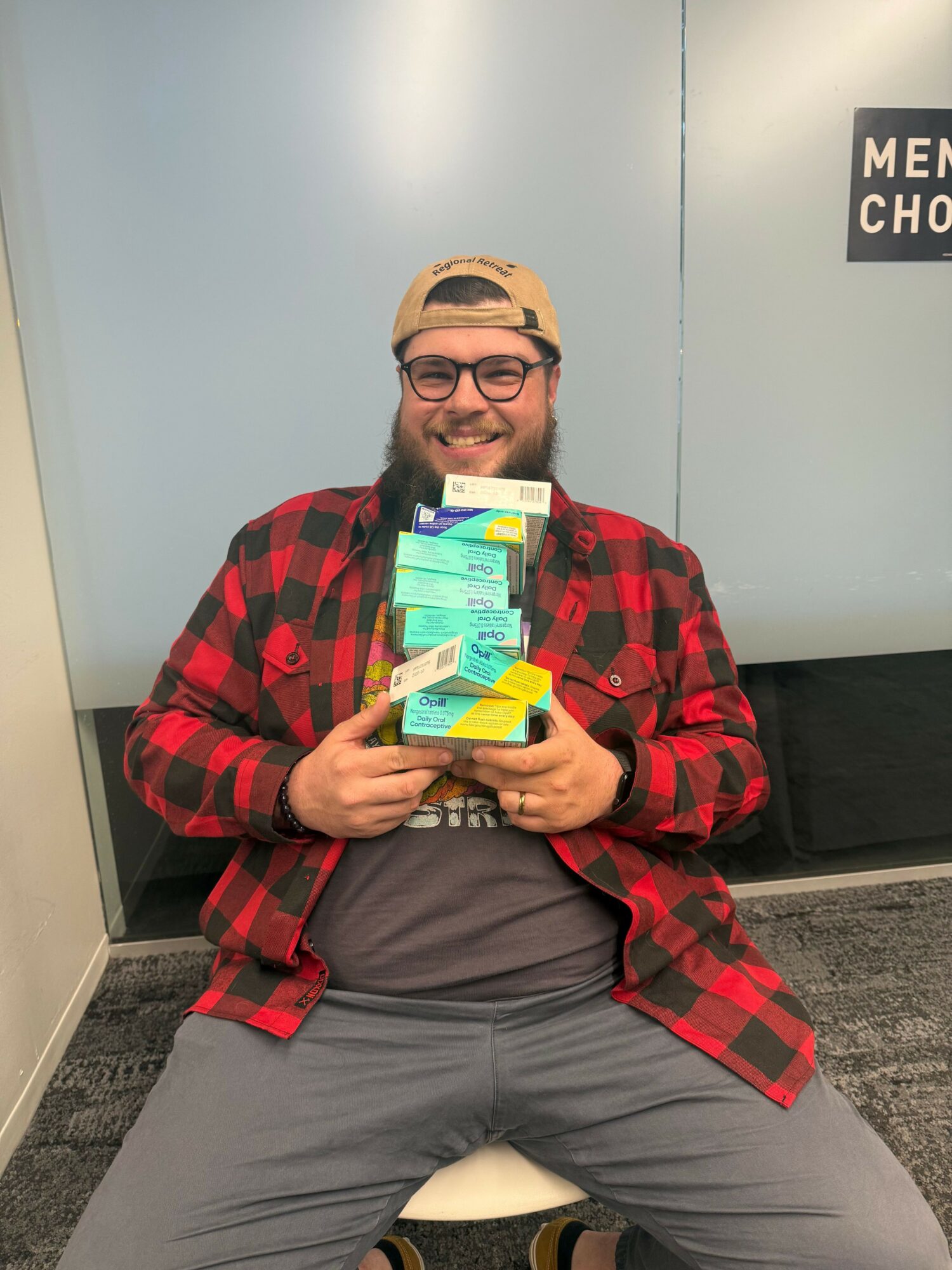 Person sitting on the floor holding multiple boxes and packages, wearing glasses, a cap, and a red plaid shirt, in a medical facility.