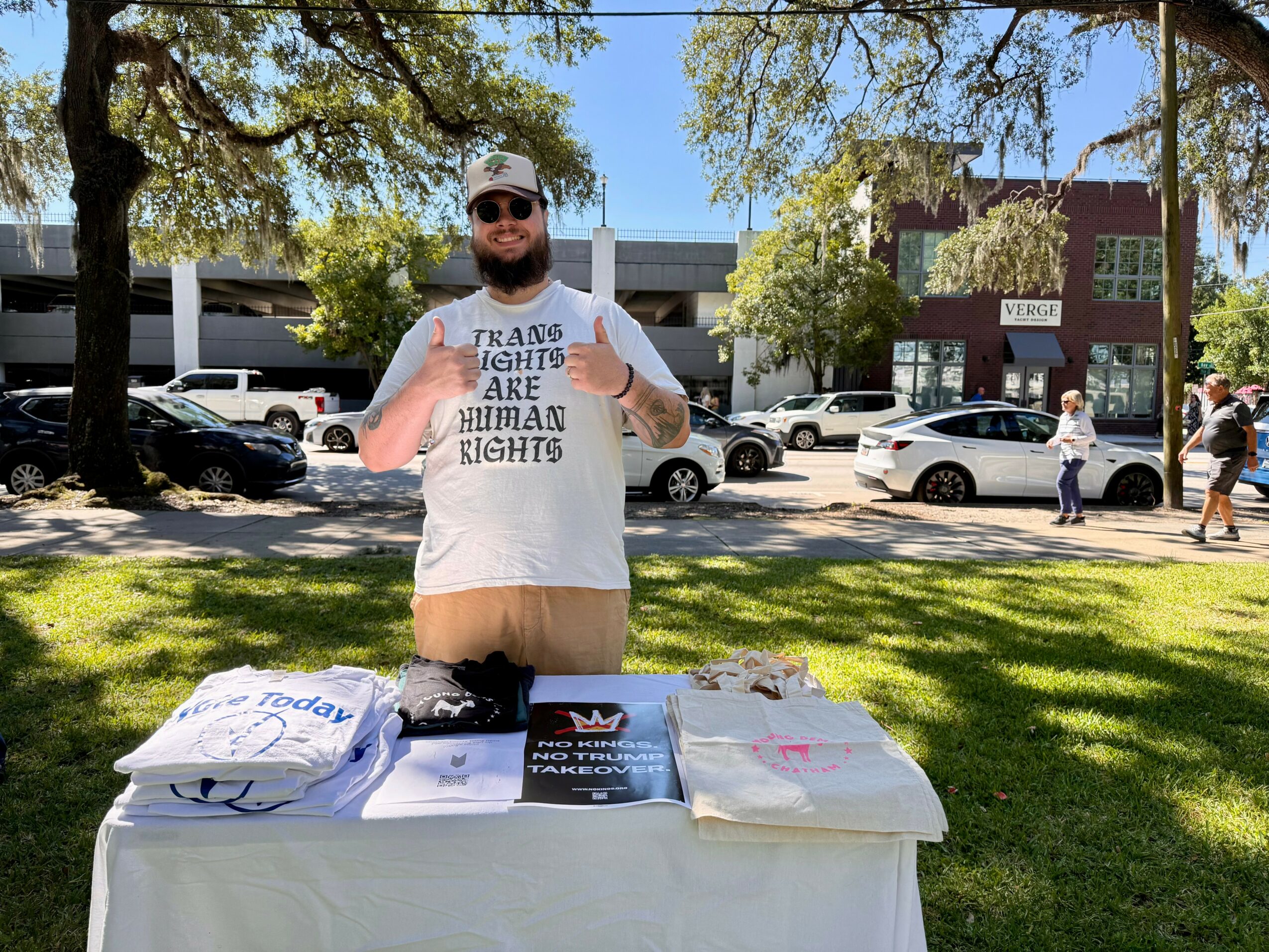 Man standing outdoors behind a table with shirts and books, holding a sign, in a park with trees and buildings in background.