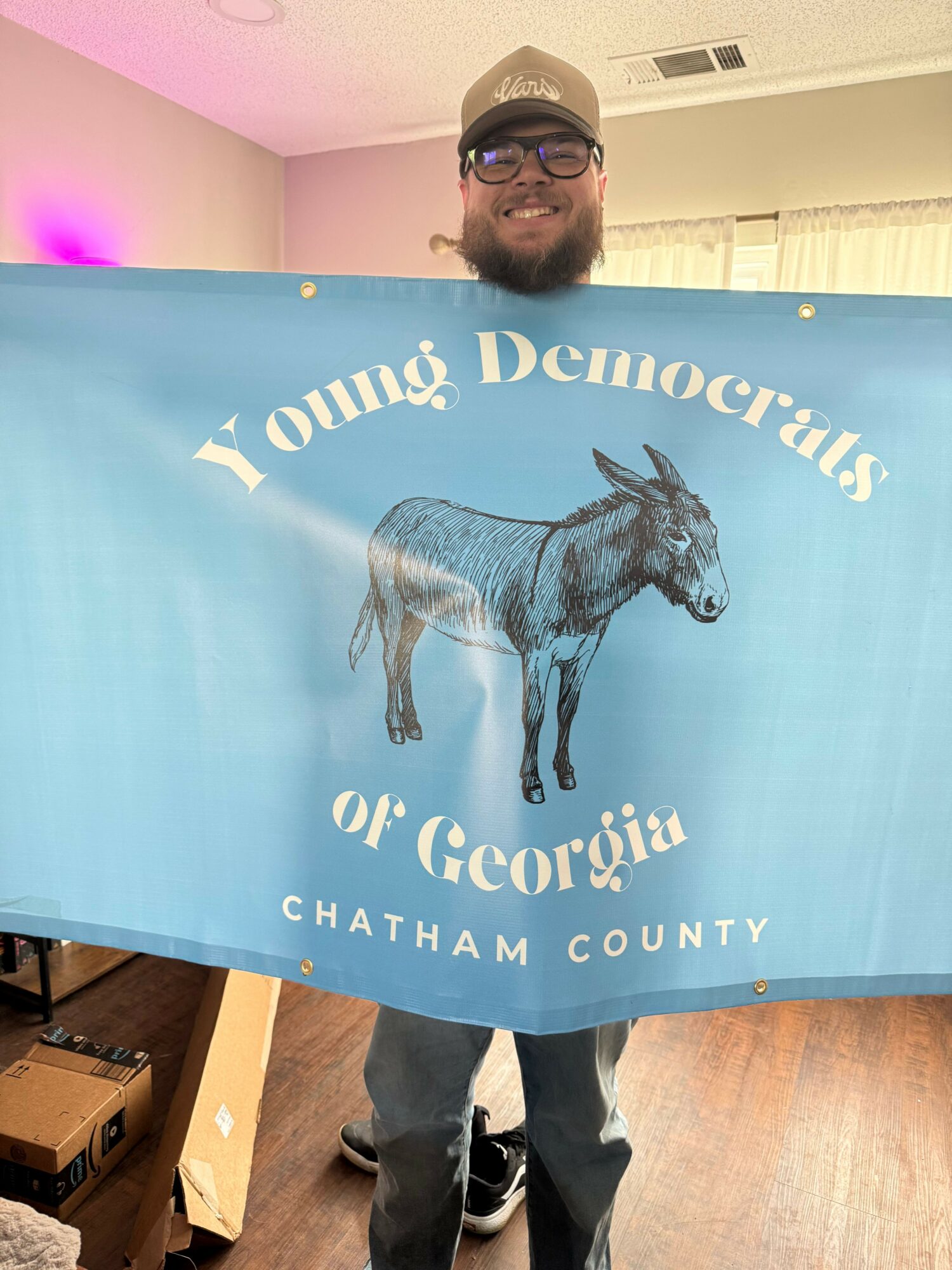 Young man standing next to a blue banner with a horse illustration and text, indoors with pink and white walls.