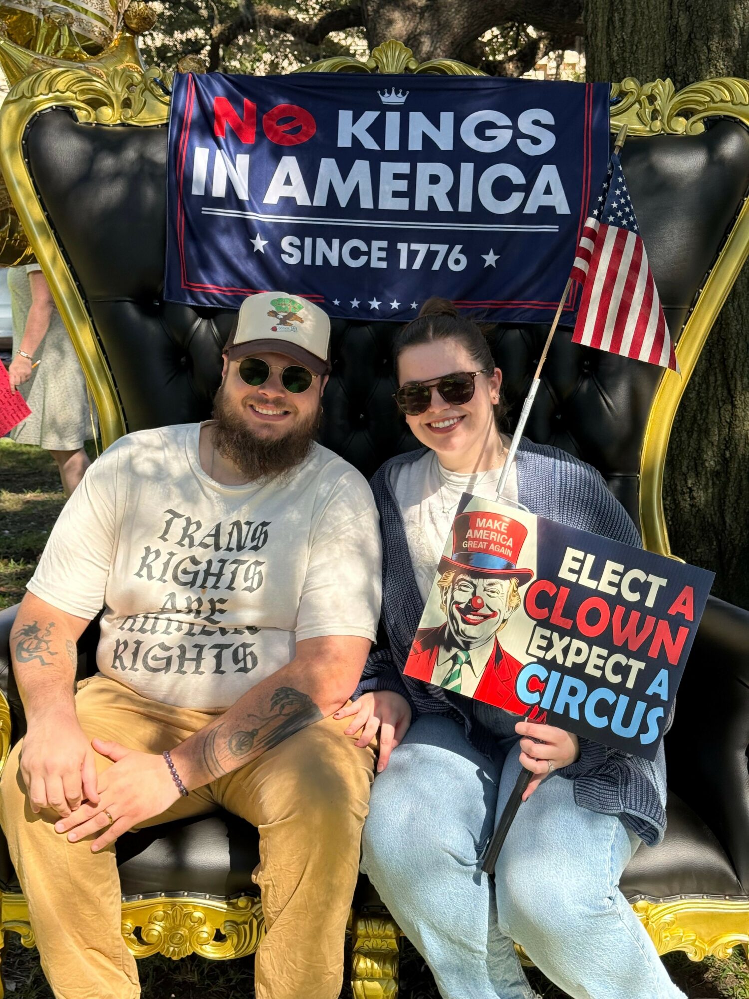 Two men sitting on a throne-like chair holding protest signs, with a flag and gold decorations around them.