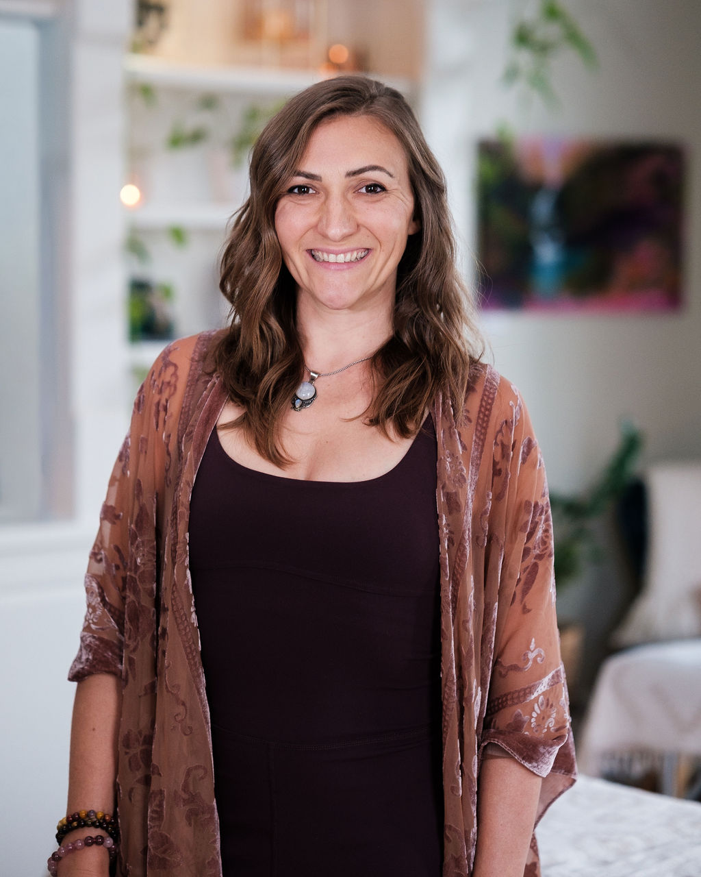Woman with wavy brown hair smiling, wearing a black top and a brown patterned shawl, in a cozy room with plants and artwork