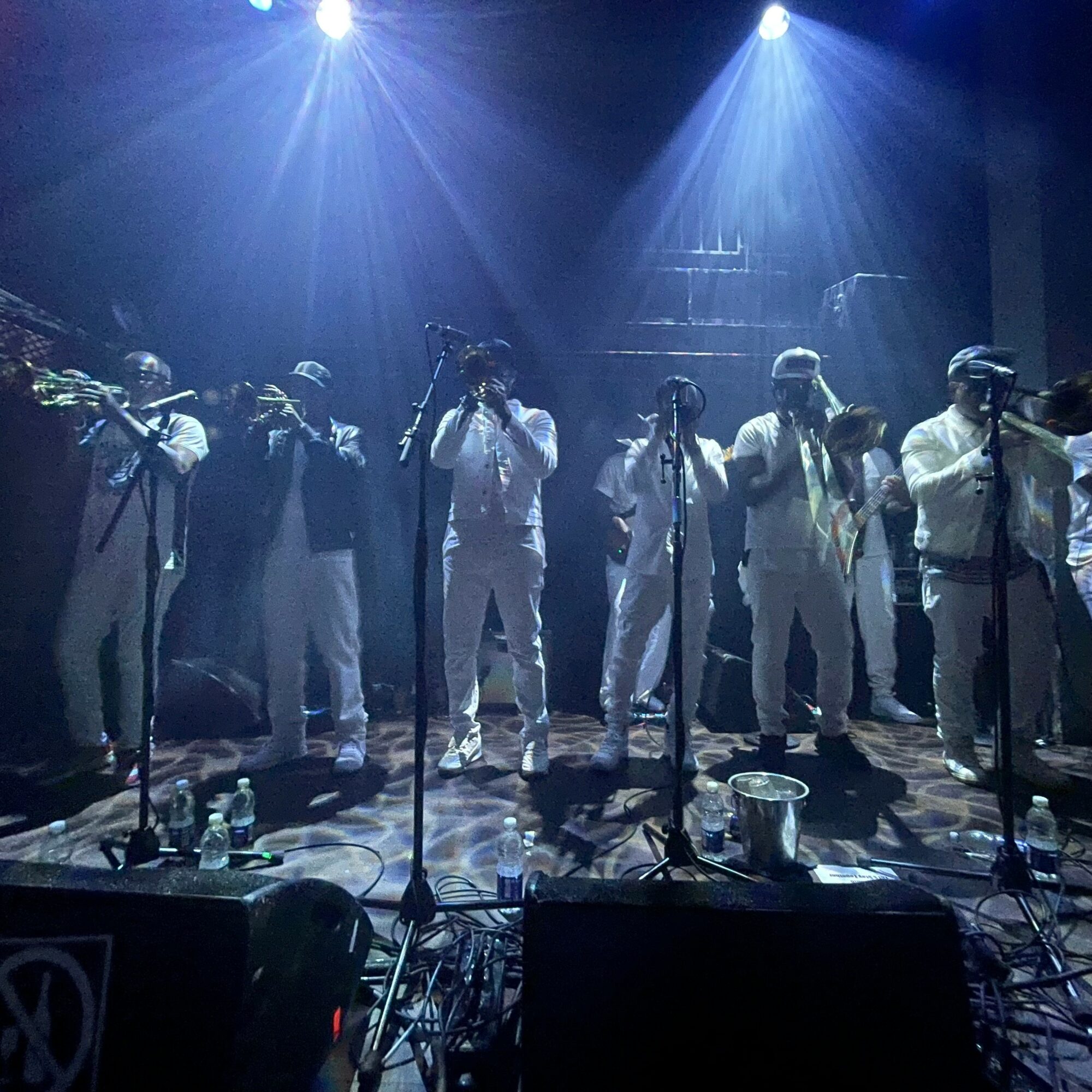 Group of people on stage with microphones, performing under blue and white lights, with equipment and water bottles in front.